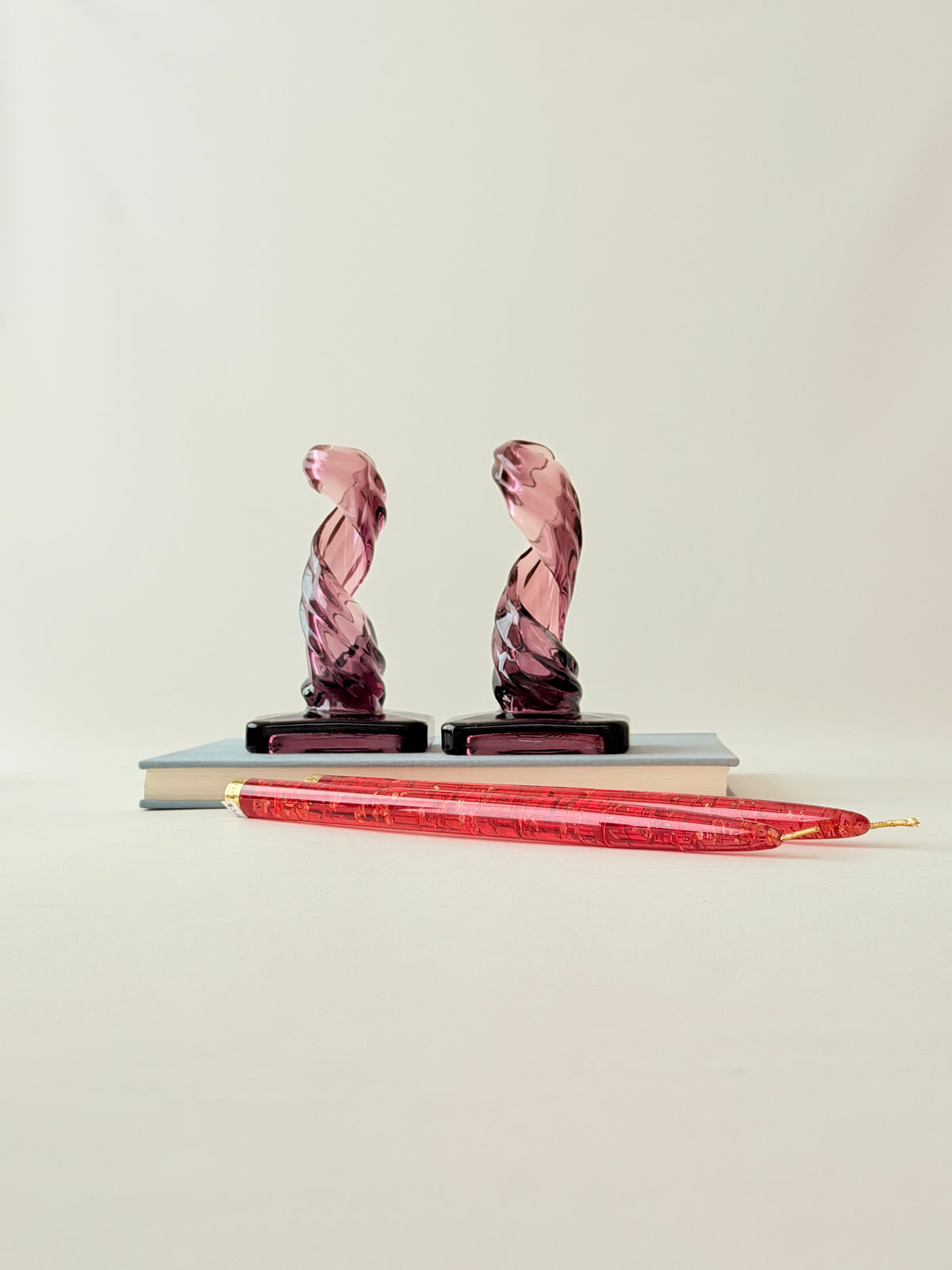 A pair of purple twisted glass candleholders and a pair of tall red lucite candles, displayed on a book against a plain background.