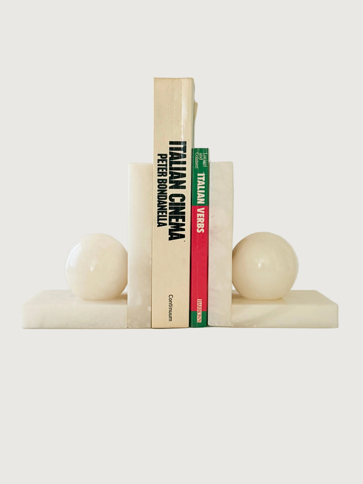 A pair of cream-colored marble bookends shaped like spheres on rectangular bases, holding up two books against a clean, light gray studio background.