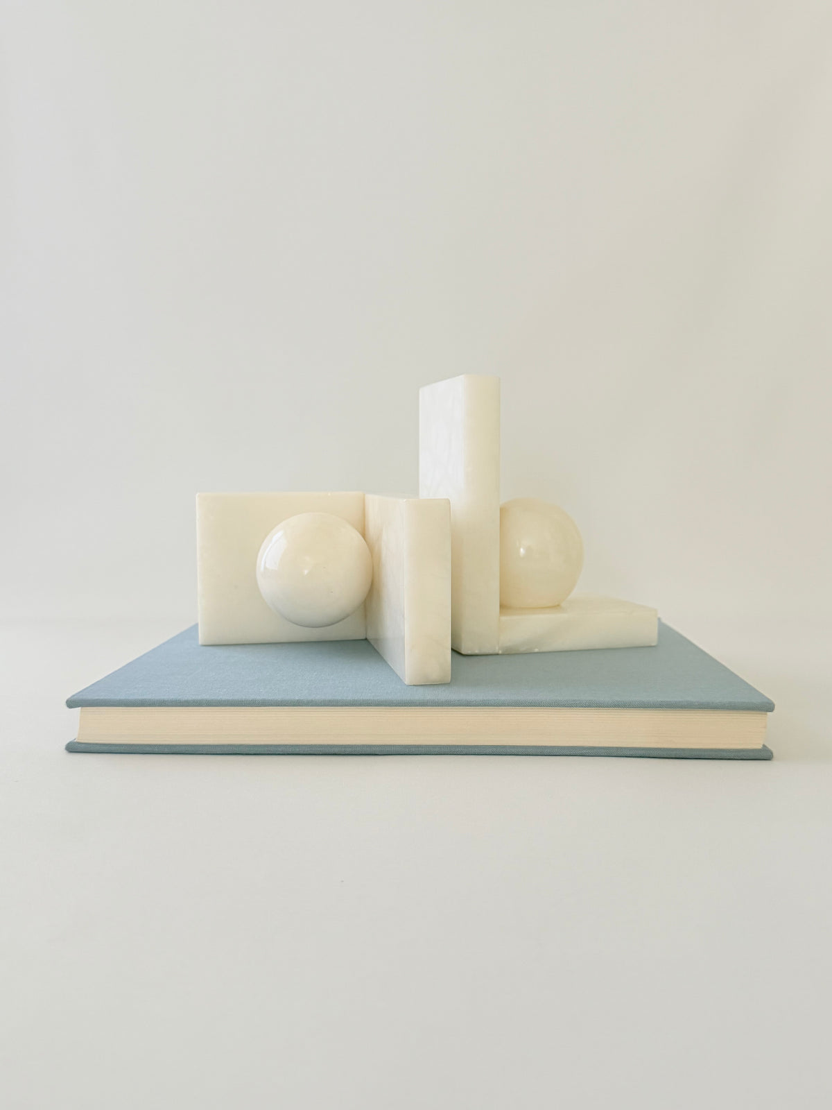 A minimalist arrangement of cream-colored geometric blocks and spheres displayed on top of a light blue hardcover book against a clean, off-white background.