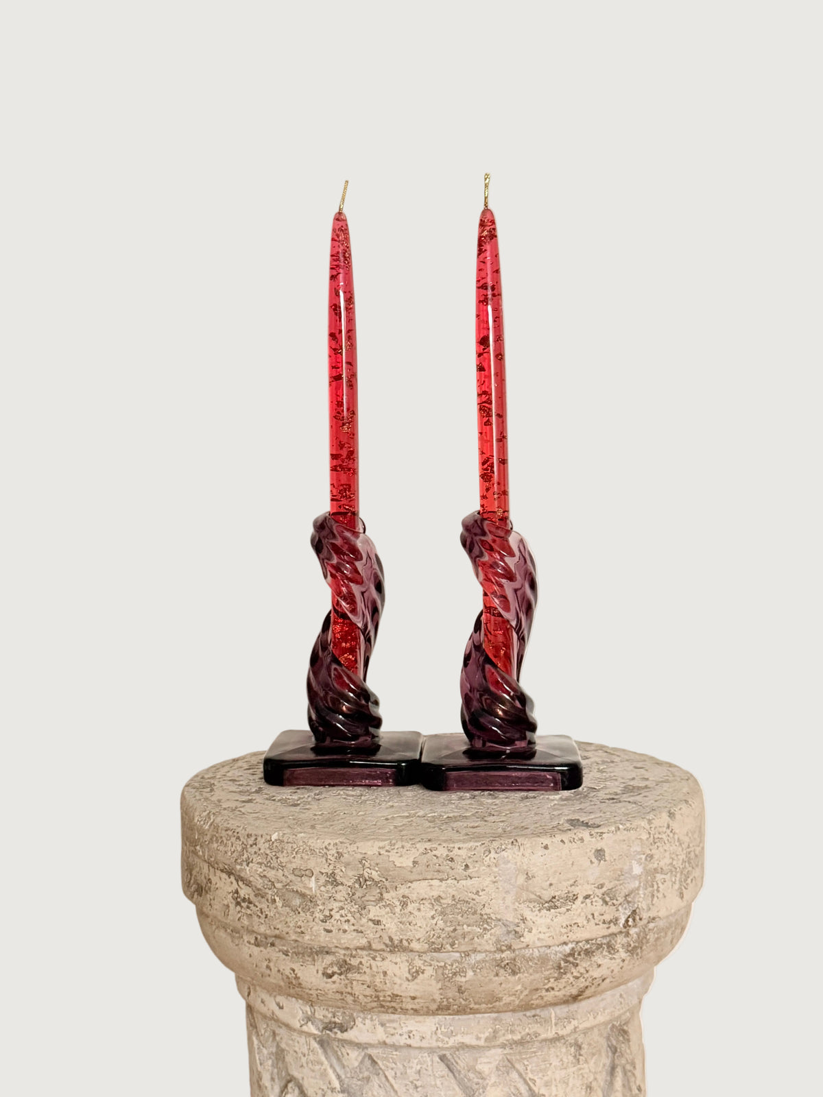 A pair of tall red lucite candles in  purple twisted glass candleholders, displayed on a stone pedestal against a plain off-white wall.