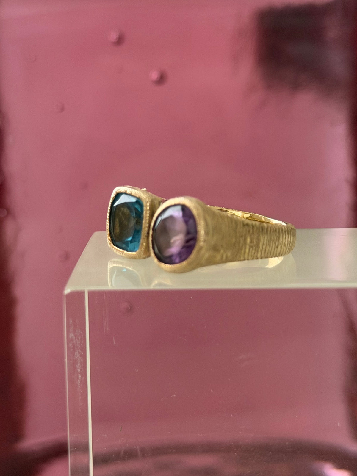 Side view of a textured gold ring with a purple and blue stone displayed on a clear acrylic block against a pink background.