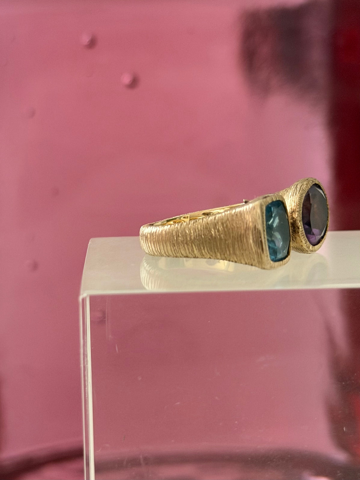 A textured vermeil ring with two stones, one blue and one dark purple, displayed on a clear acrylic block against a pink background.