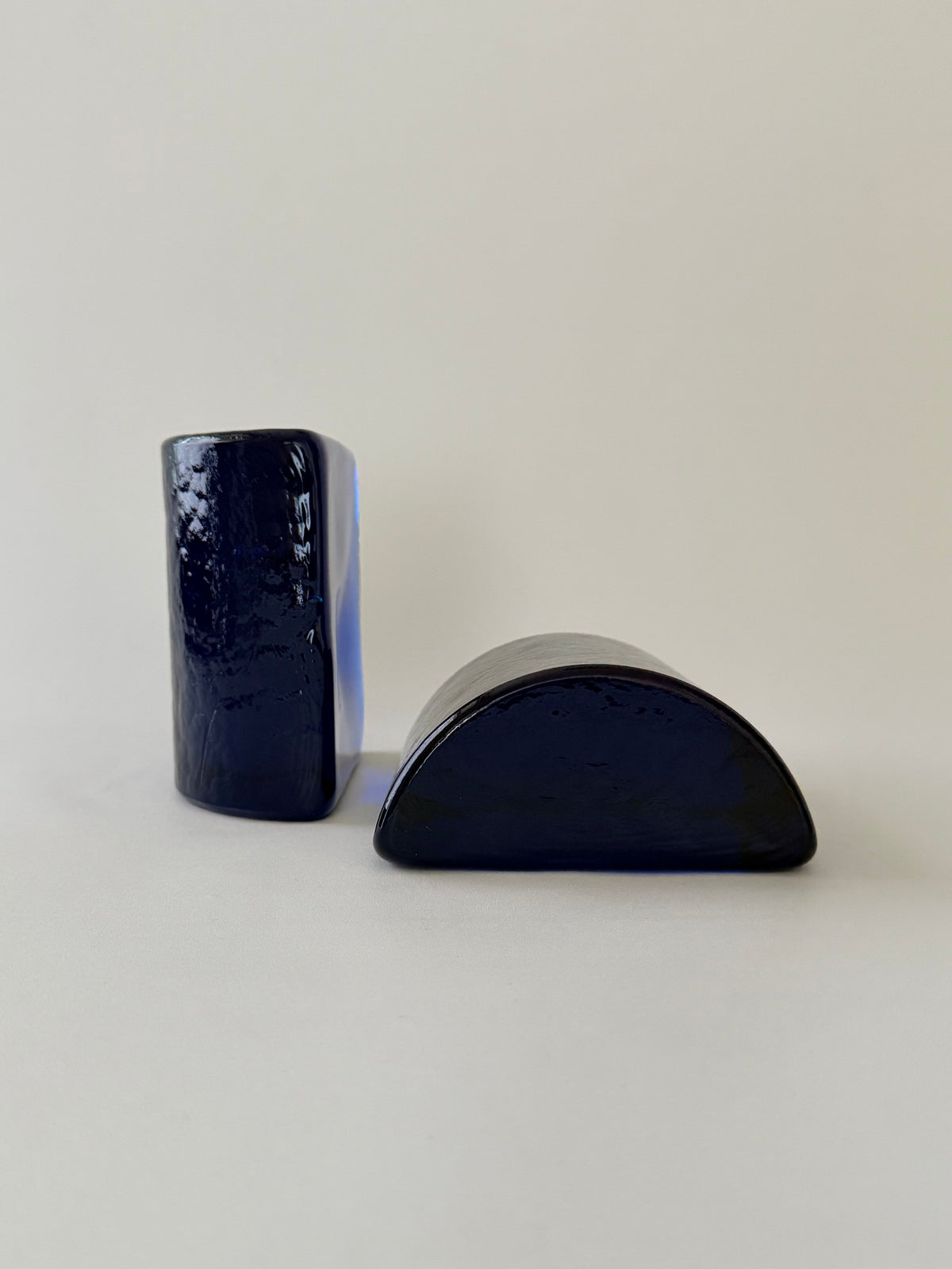 A pair of cobalt blue glass half moon bookends, one standing, the other face down on a plain background.
