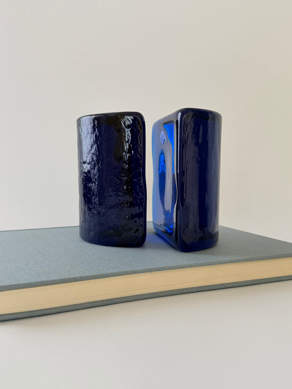 Two cobalt blue glass bookends displayed on a gray hardcover book against a plain off-white background.