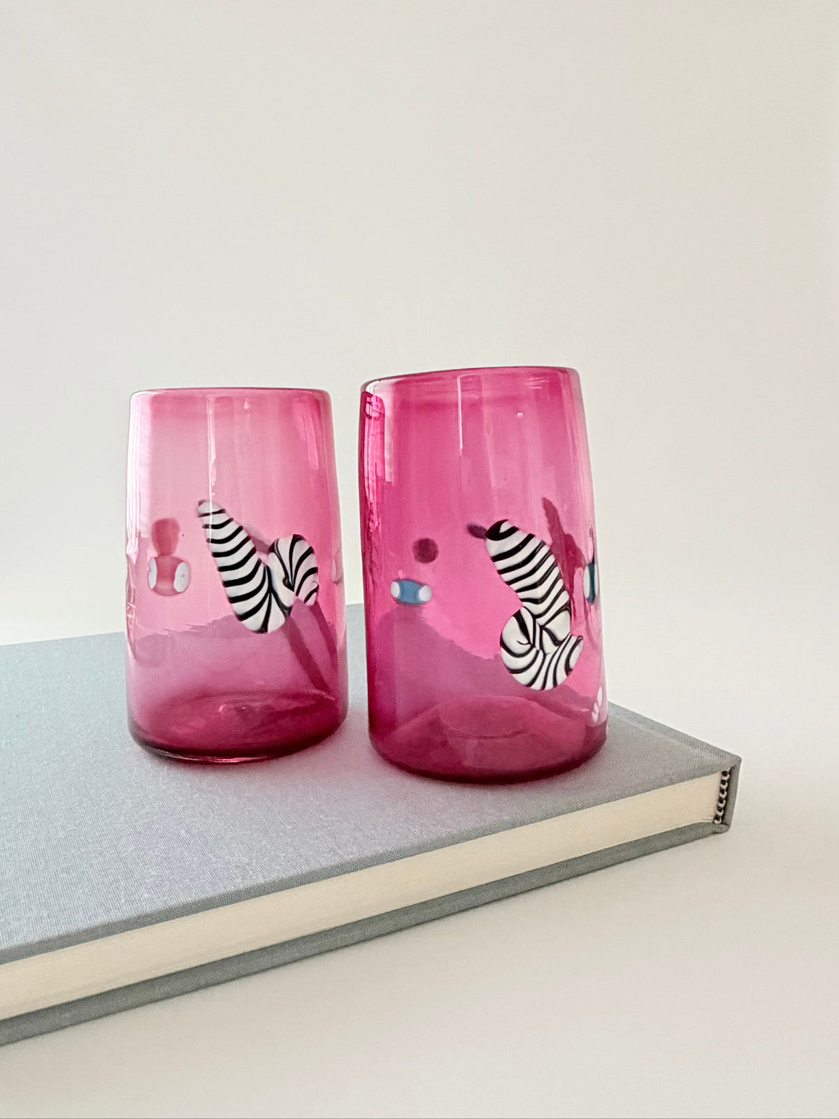 A pair of pink glass tumblers with black and white zebra print and small colorful decorative details on book.