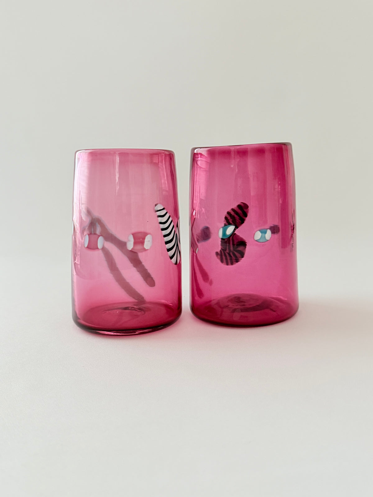 A pair of pink glass tumblers with colorful decorative details against a light gray background.