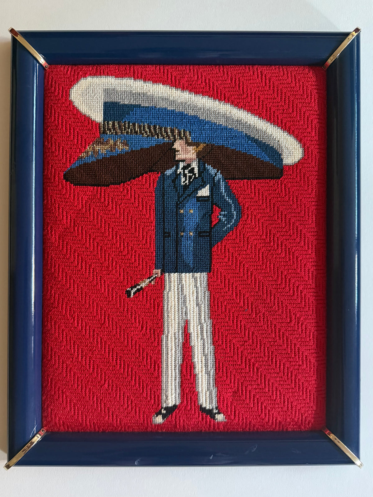 A framed needlepoint artwork featuring a stylized  yacht man in a blue suit and white striped trousers with an oversized  blue, white, and brown hat, set against a textured red background. The piece is enclosed in a dark blue frame with gold-toned corner accents.