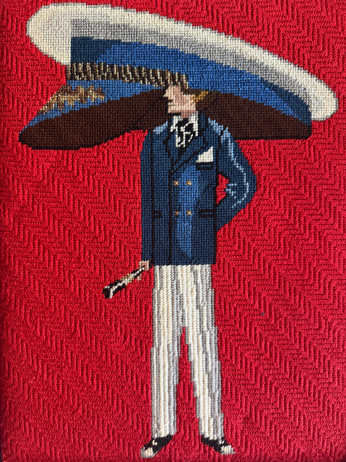 A close-up of a woven needlepoint design featuring a yacht man in a blue double-breasted suit, white striped trousers, and a large blue and white hat, set against a textured red background.