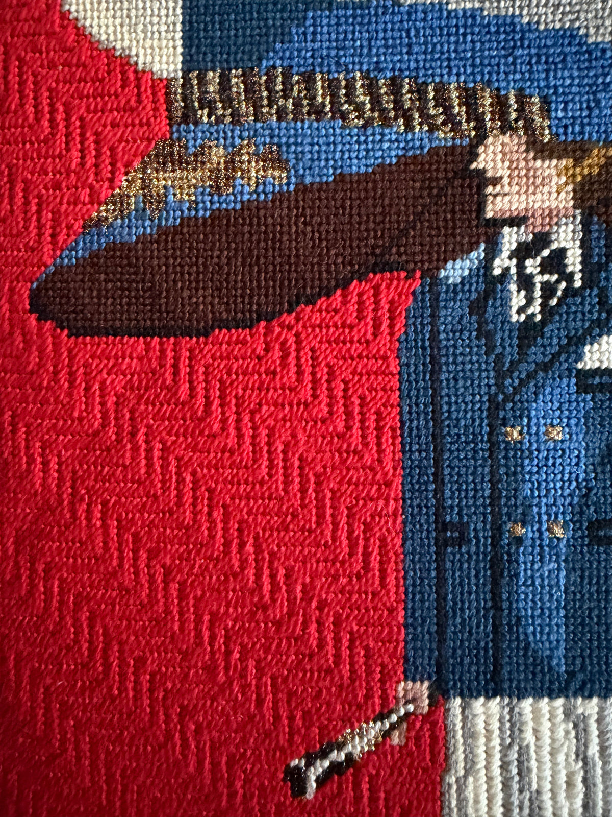 Close-up of a woven needlepoint showing part of a person in a blue suit and hat with a large red field, with an oversized brown hat brim, white shirt, and gold buttons.