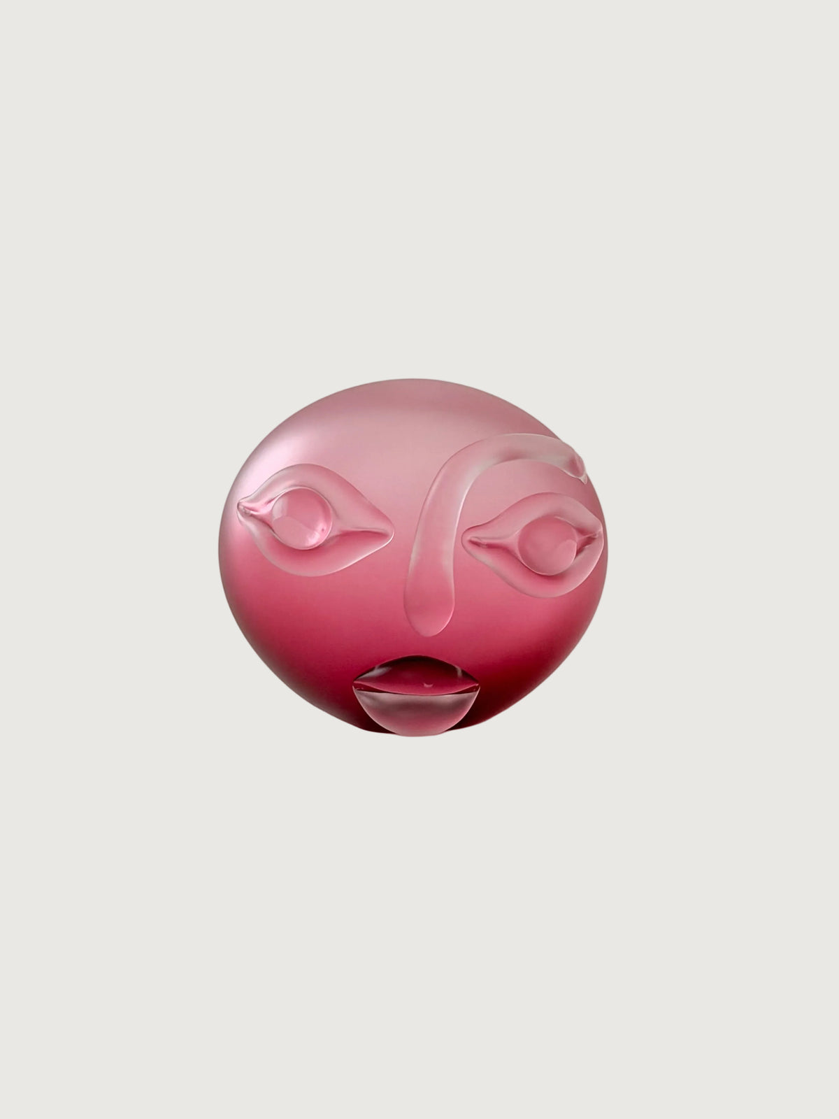 A pink frosted glass sculpture shaped like a stylized face, photographed against a neutral light gray background.