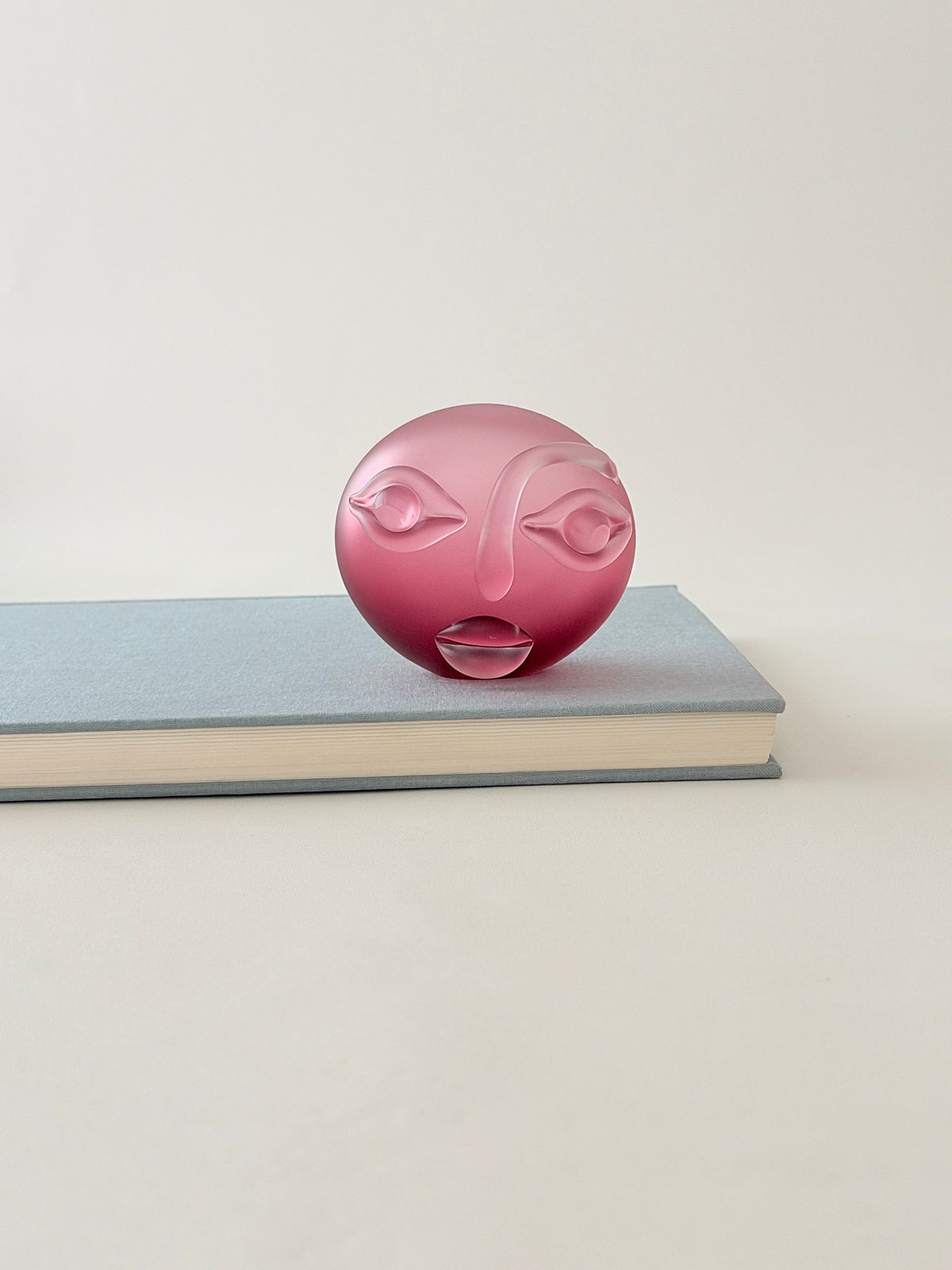 A pink glass sculpture of a stylized face with resting on top of a light blue hardcover book against a neutral off-white background.