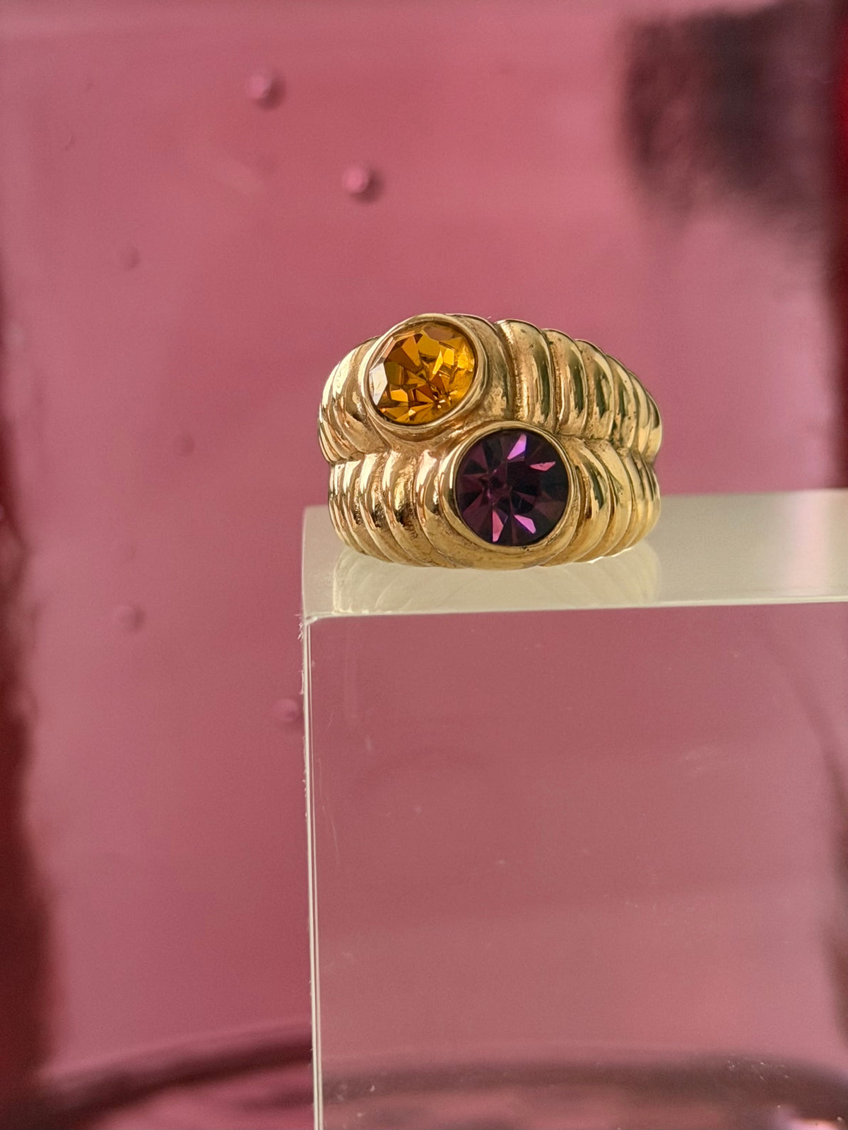 A gold ring with two round-cut gemstones, one orange and one purple, displayed on a clear acrylic block against a pink background.