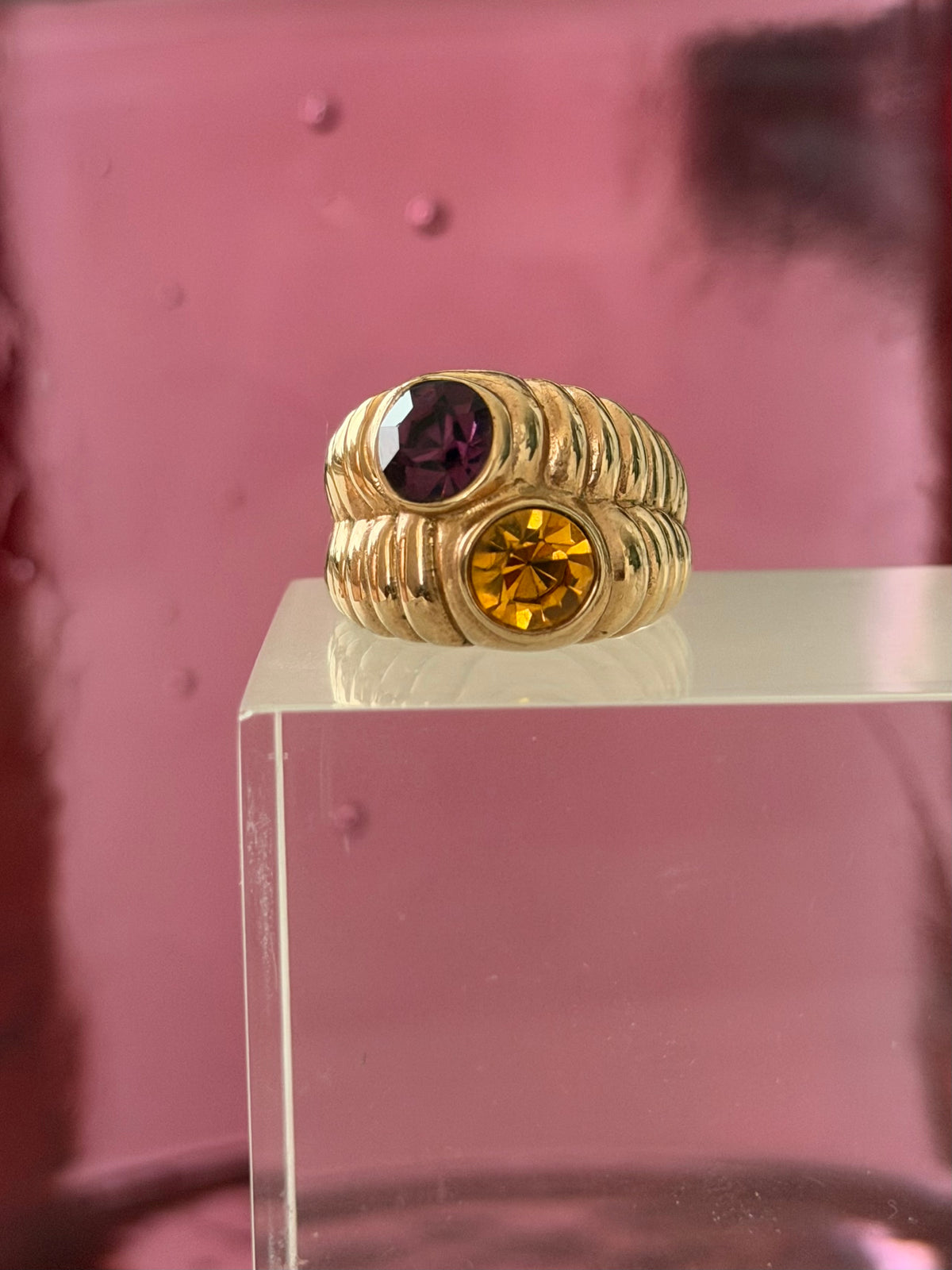A gold ring with a textured, ribbed band featuring a large round yellow gemstone and a smaller oval purple gemstone, displayed on a clear acrylic block against a pink background.