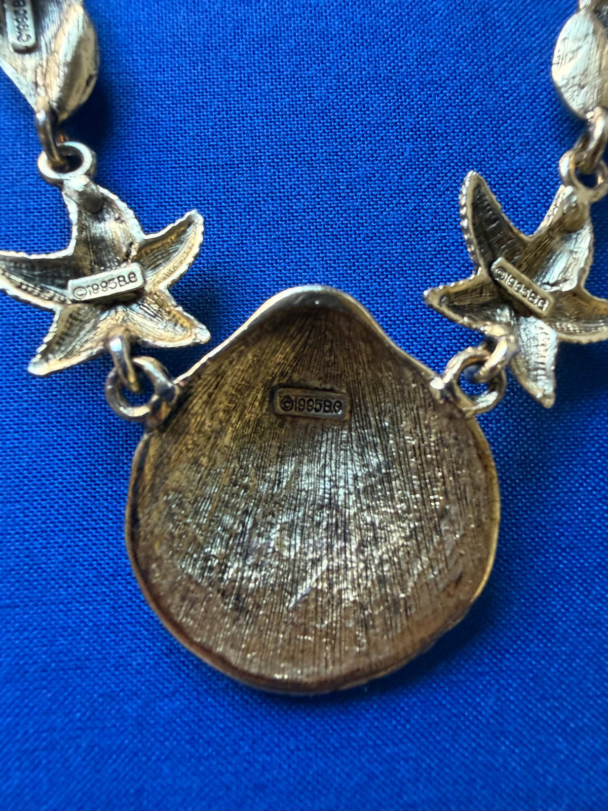 Close-up of a gold-toned necklace featuring the back of a shell and starfish signed 1995 BG on a vibrant blue textured background.
