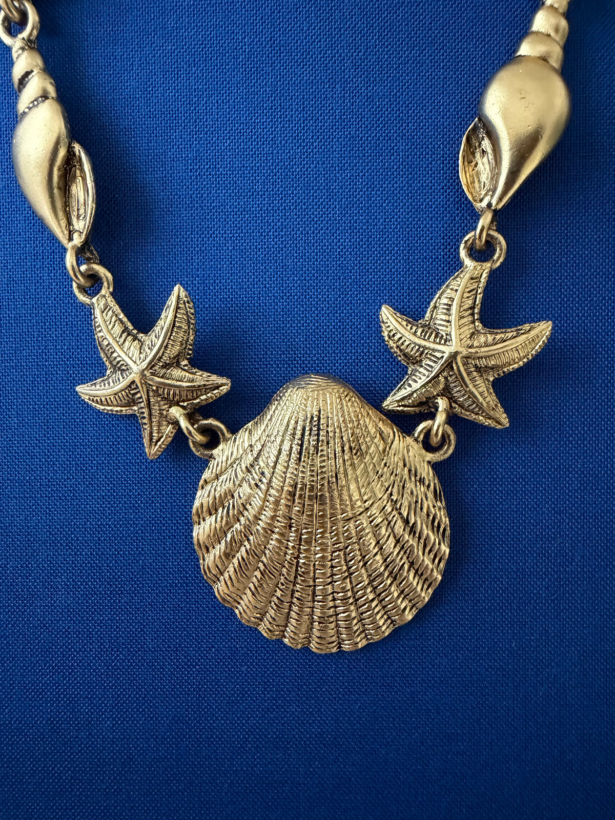 Gold-toned shell necklace featuring a large textured scallop shell pendant, two starfish charms, and spiral shell links, displayed on a blue book.
