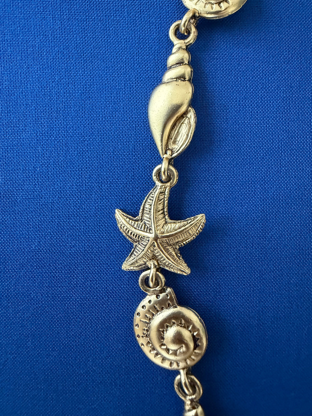 A close-up of a gold-toned jewelry charm featuring a starfish, a spiral seashell, and a smaller shell charm, laid against a blue book.