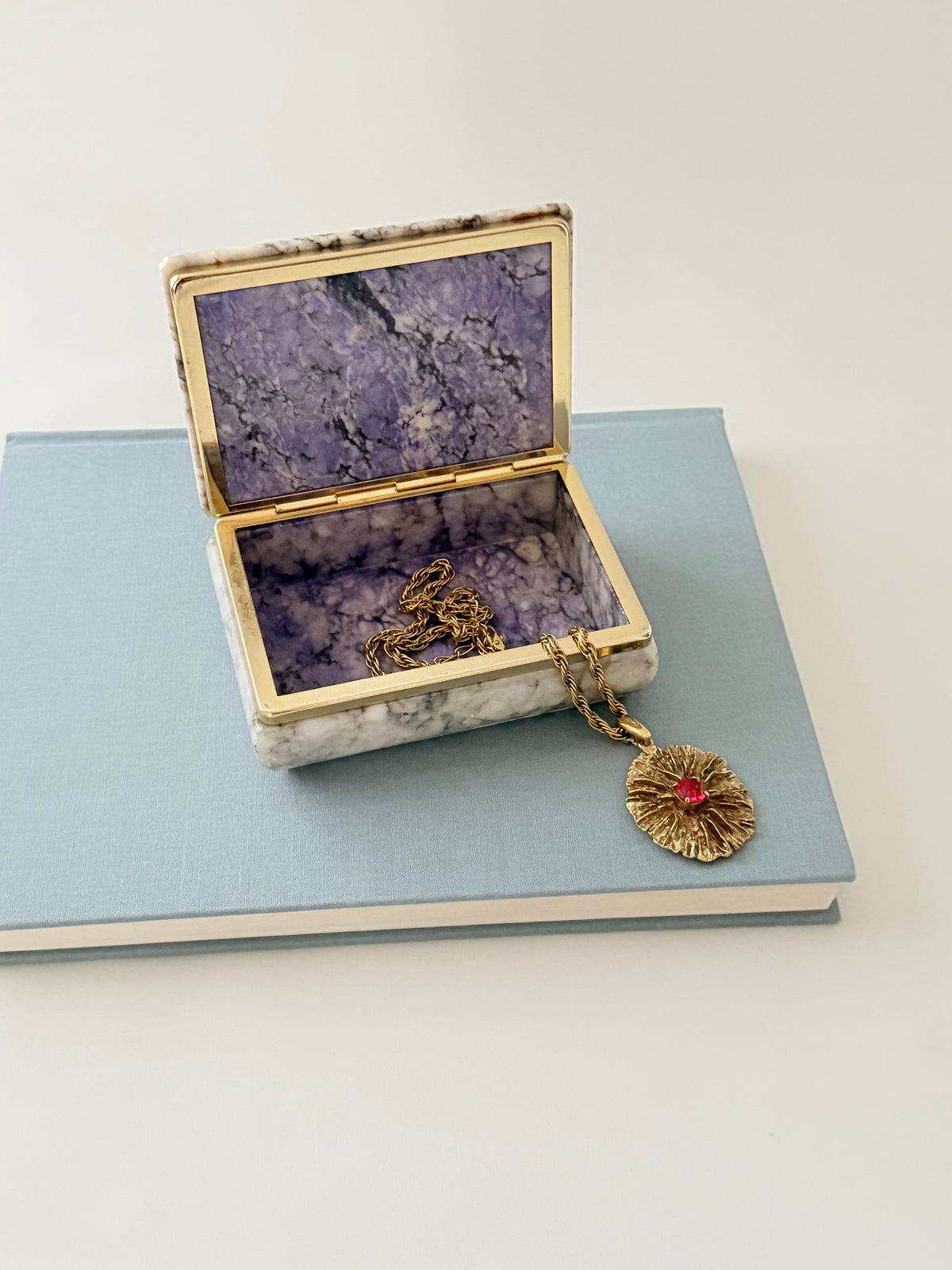 Vintage gray hand-carved 1970s Italian alabaster box with  purple interior styled with a gold-toned YSL necklace on a light blue book.
