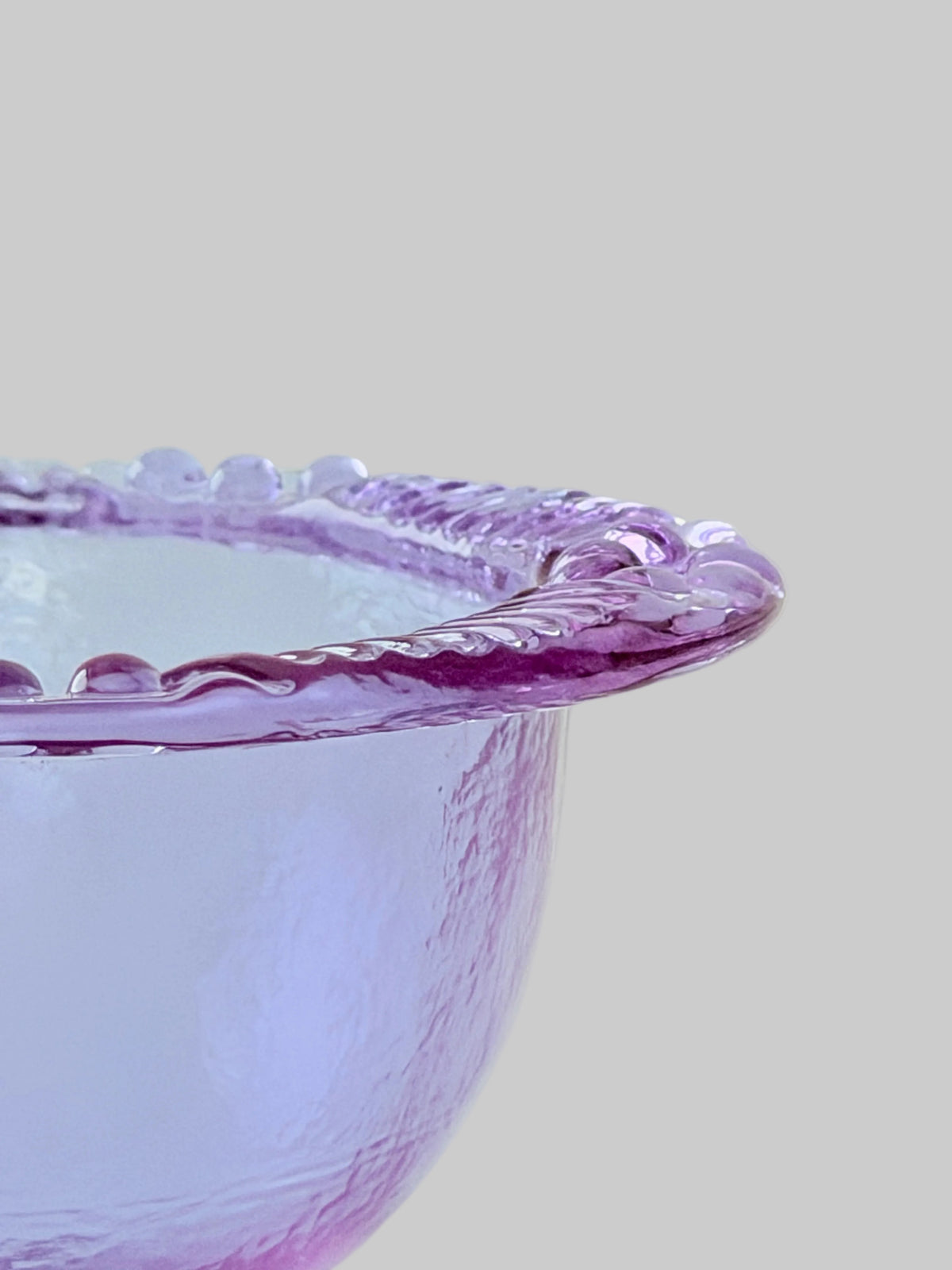 A close-up of a translucent lavender-purple blush glass bowl with a decorative wavy rim, set against a soft, pale cream background.