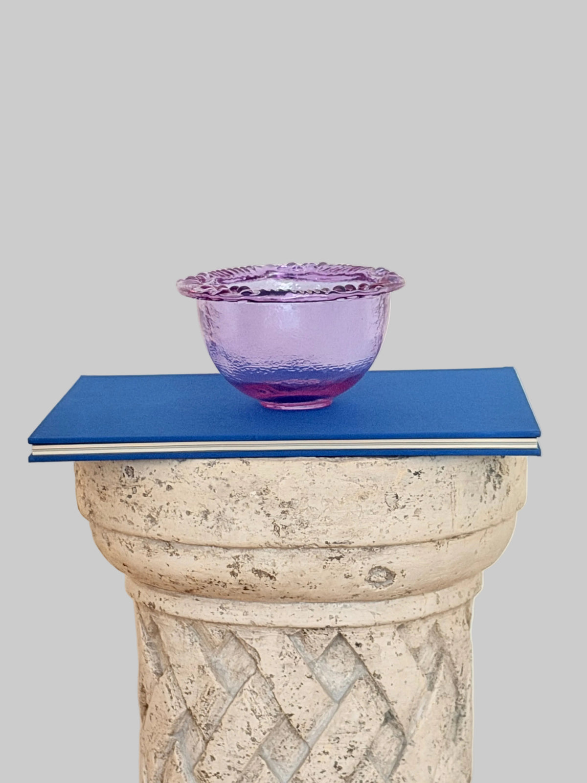 A textured pink purple blush glass bowl with a scalloped rim n a blue book placed on top of a stone pedestal against a plain off-white wall.