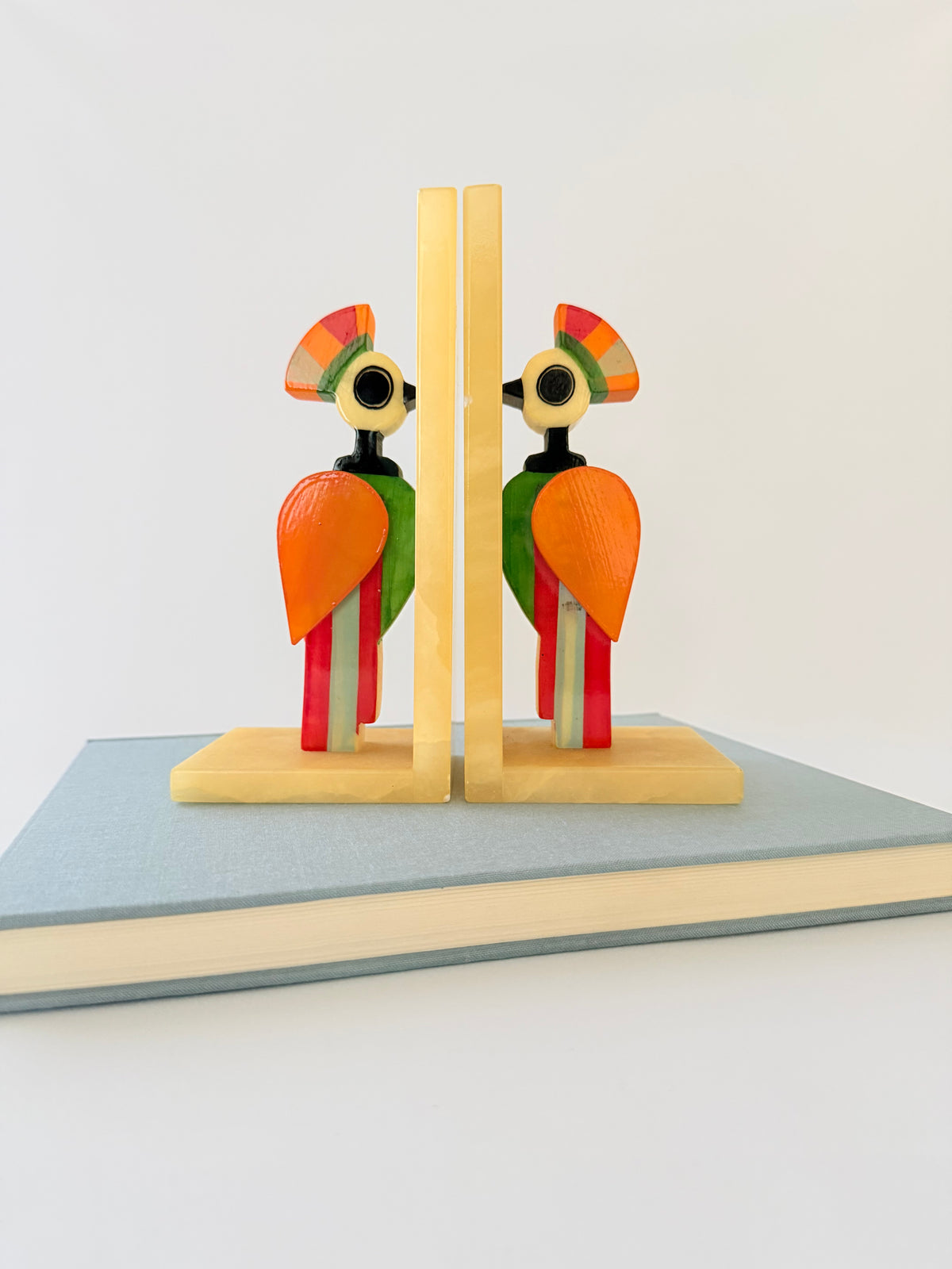 A pair of colorful alabaster bookends shaped like stylized birds set against a plain white background and on a book. Home Decor. Vintage. Collectible. 