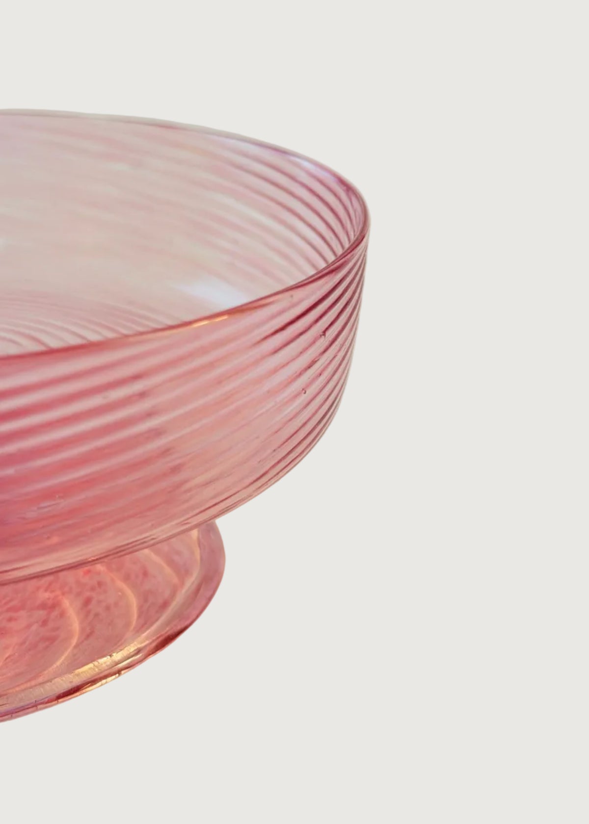 A close-up of a pink ribbed glass bowl with a pedestal base, set against a solid cream-colored background.