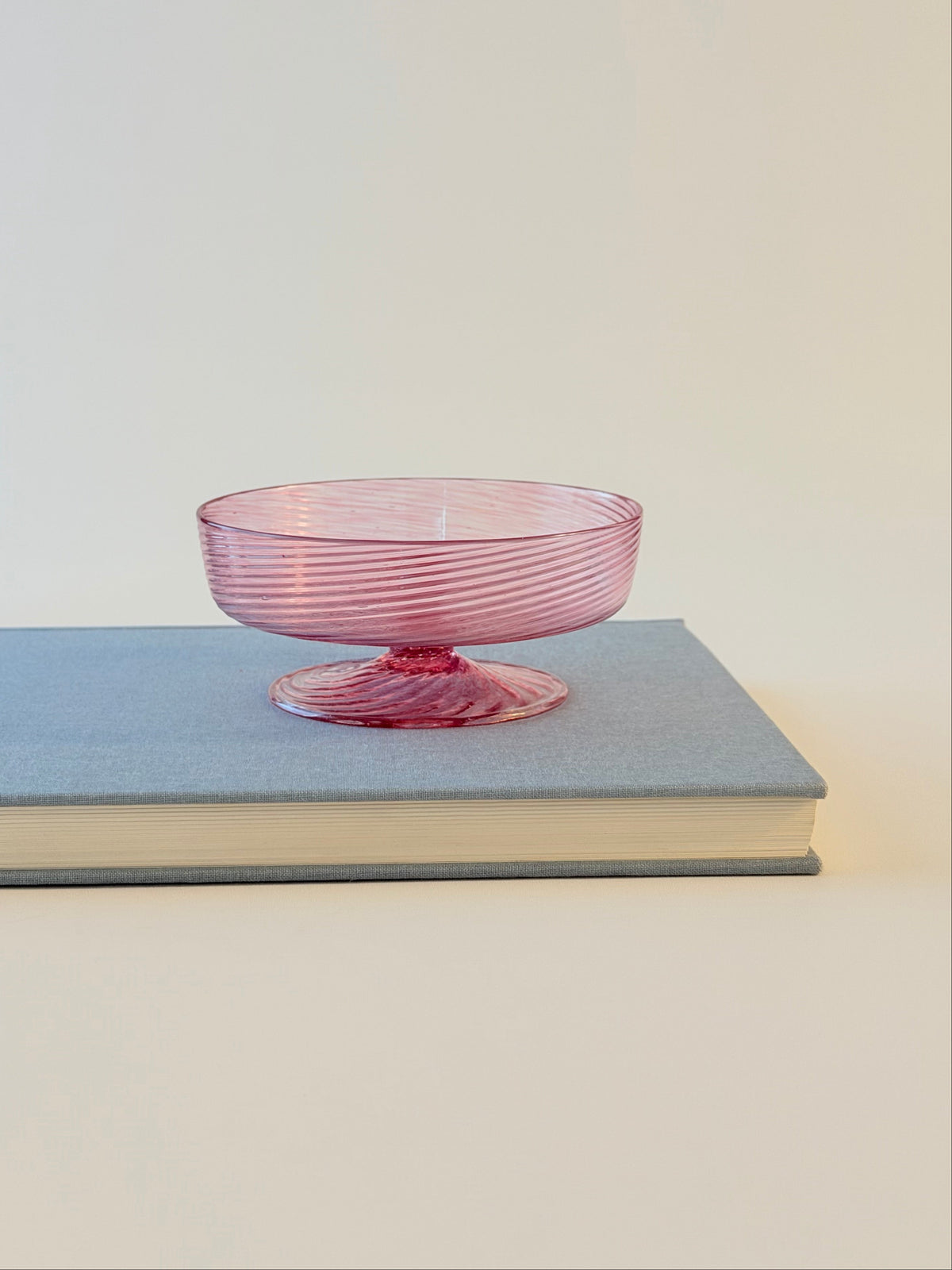 A pink ribbed glass pedestal bowl resting on top of a light blue hardcover book against a plain off-white background.