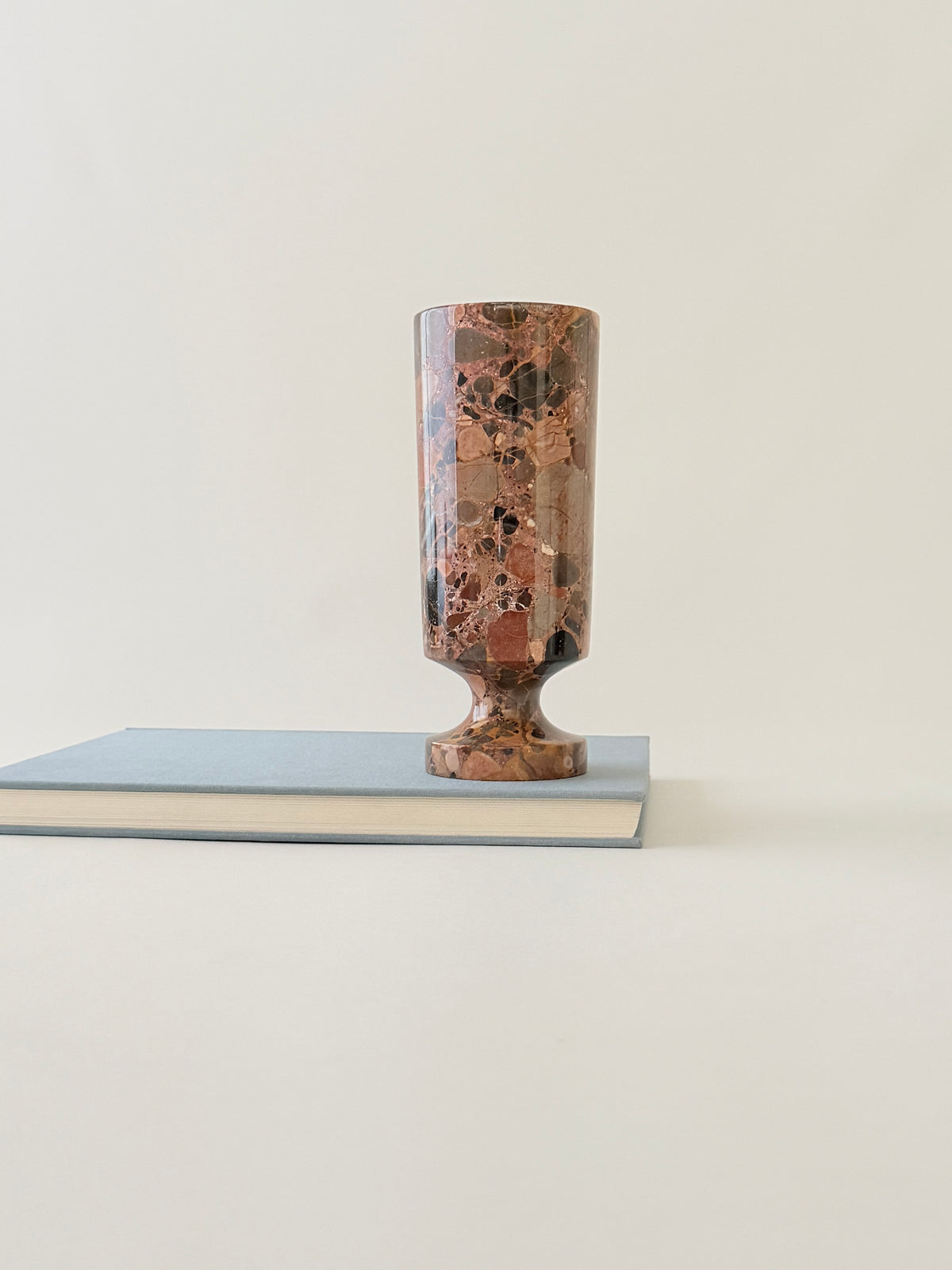 A breccia marble cylindrical vase  with a natural earth-toned stone featuring a fragmented, multicolored "brecciated" look and veining on a book