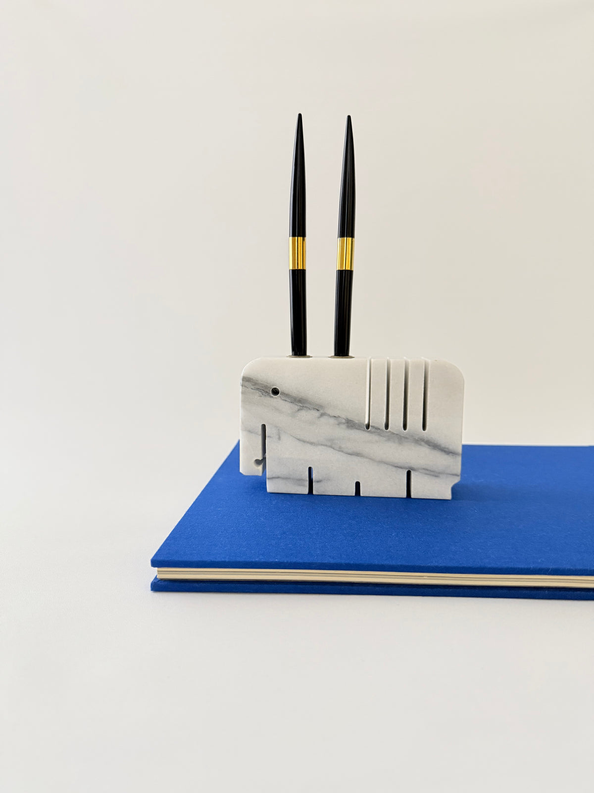 Two pens in a white and gray marble elephant penholder with slots for two pens and four slots for notes on a blue book.