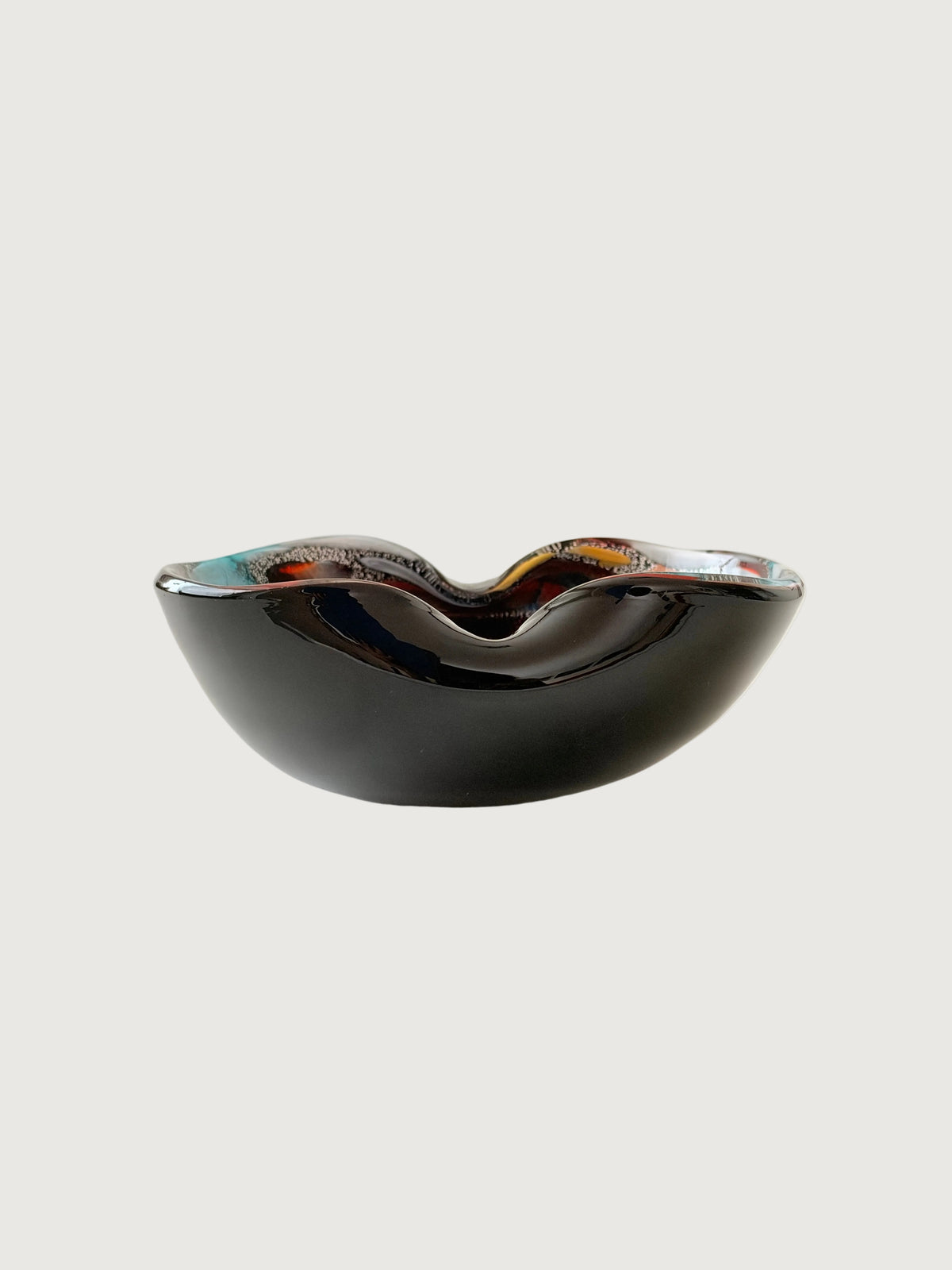 Side view  of a striking mid-century hand-formed Murano "Tutti Frutti" bowl with two soft folds on thick black glass, a form used for bowls and ashtrays. Inside are bold colored fragments, cane sections on silver leaf and aventurine, copper crystals that make the glass sparkle.