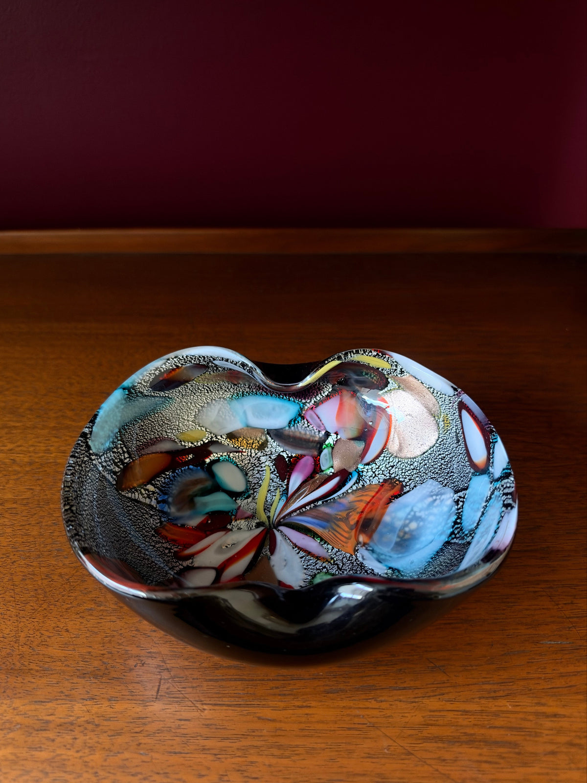 On a wood console sits a striking mid-century hand-formed Murano "Tutti Frutti" bowl with two soft folds on thick black glass, a form used for bowls and ashtrays. Inside are bold colored fragments, cane sections on silver leaf and aventurine, copper crystals that make the glass sparkle.