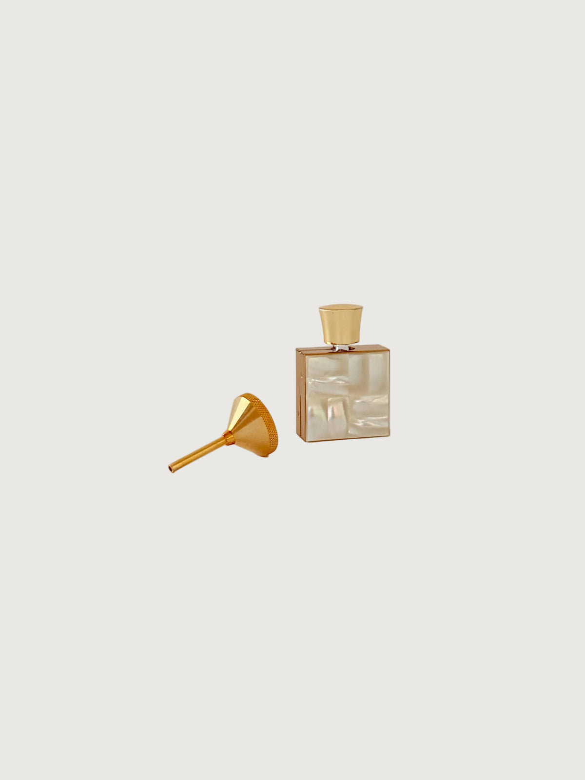 A small rectangular mother of pearl perfume bottle with a gold cap and a matching gold funnel placed beside it on a clean, light beige background.
