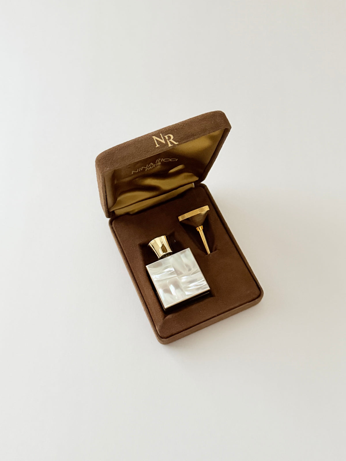 A small perfume bottle made of mother-of-pearl and gold-toned hardware, presented in an open brown velvet box with a gold monogrammed lid on a light gray background.