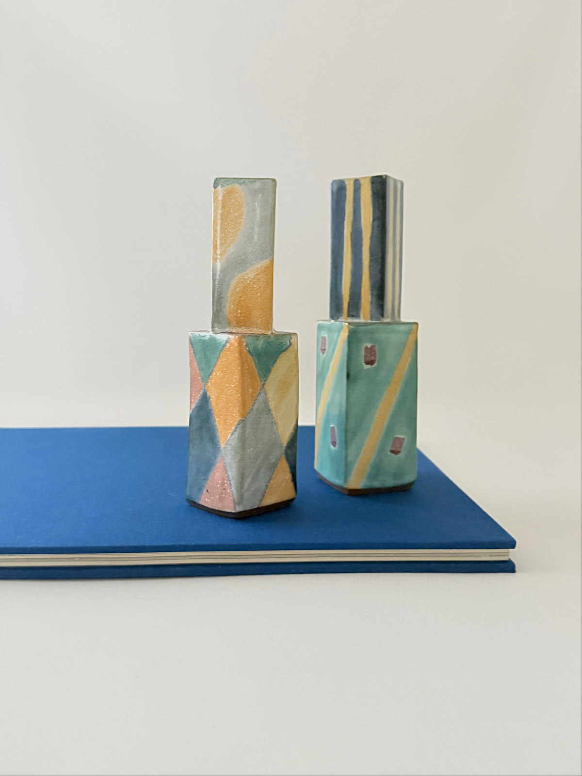 Two ceramic candleholders with abstract painted patterns in teal, yellow, orange, and gray, displayed on top of a blue book against a white background.