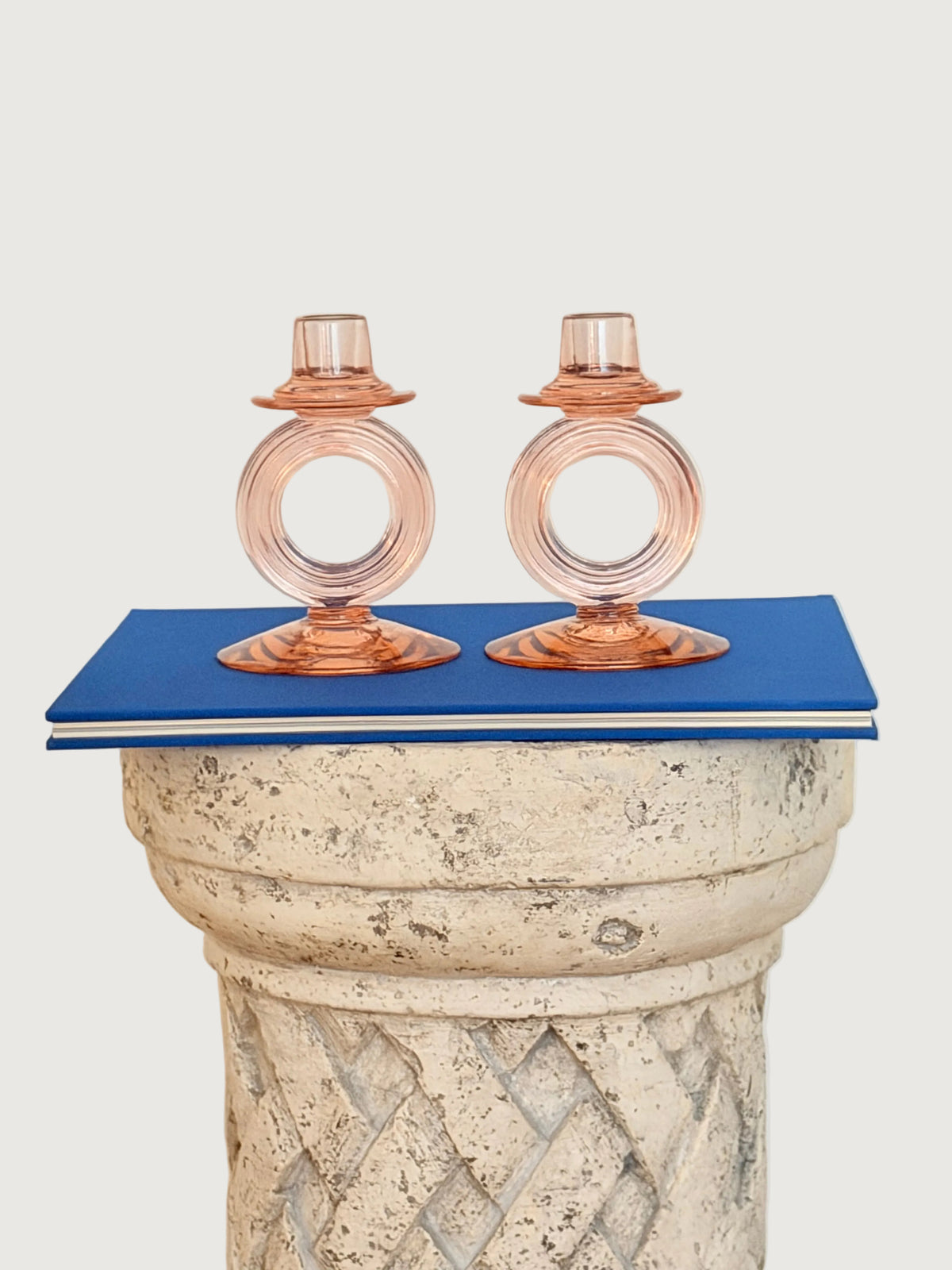 Two pink glass candle holders with circular ring details, displayed on a blue book resting on a textured stone pedestal against a plain off-white wall.