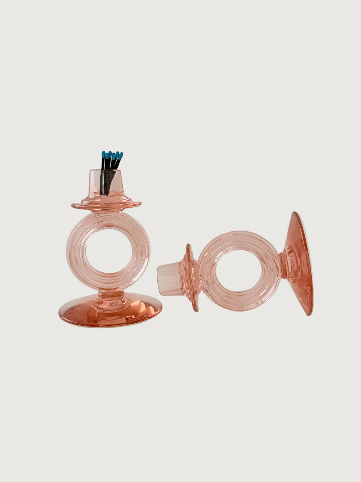 A pair of pink glass candleholders set on a light beige background, featuring a holder with a small top cup containing blue and black sticks, and a separate matching ring-shaped piece lying beside it.