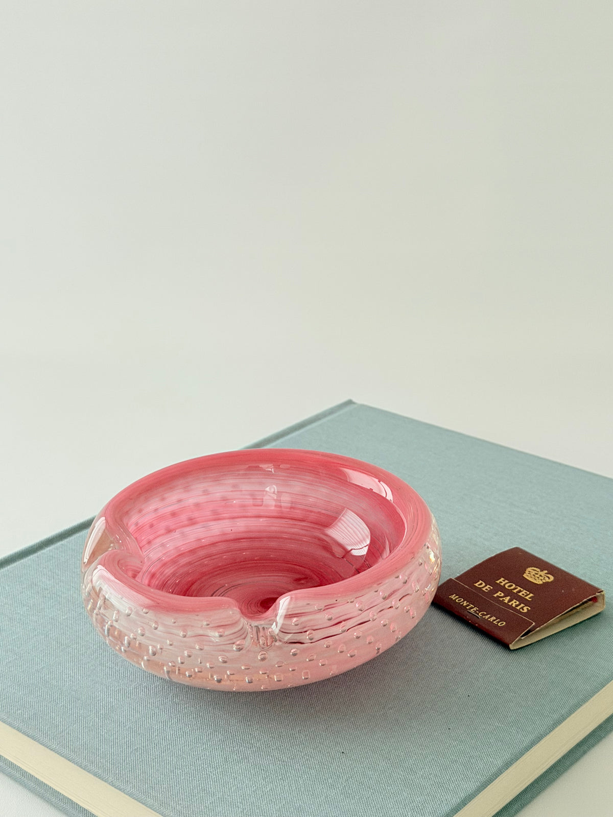 A  round vibrant pink glass ashtray with a textured, bubble-filled design, on a blue book with matches. Home Decor. Vintage. Collectible. 