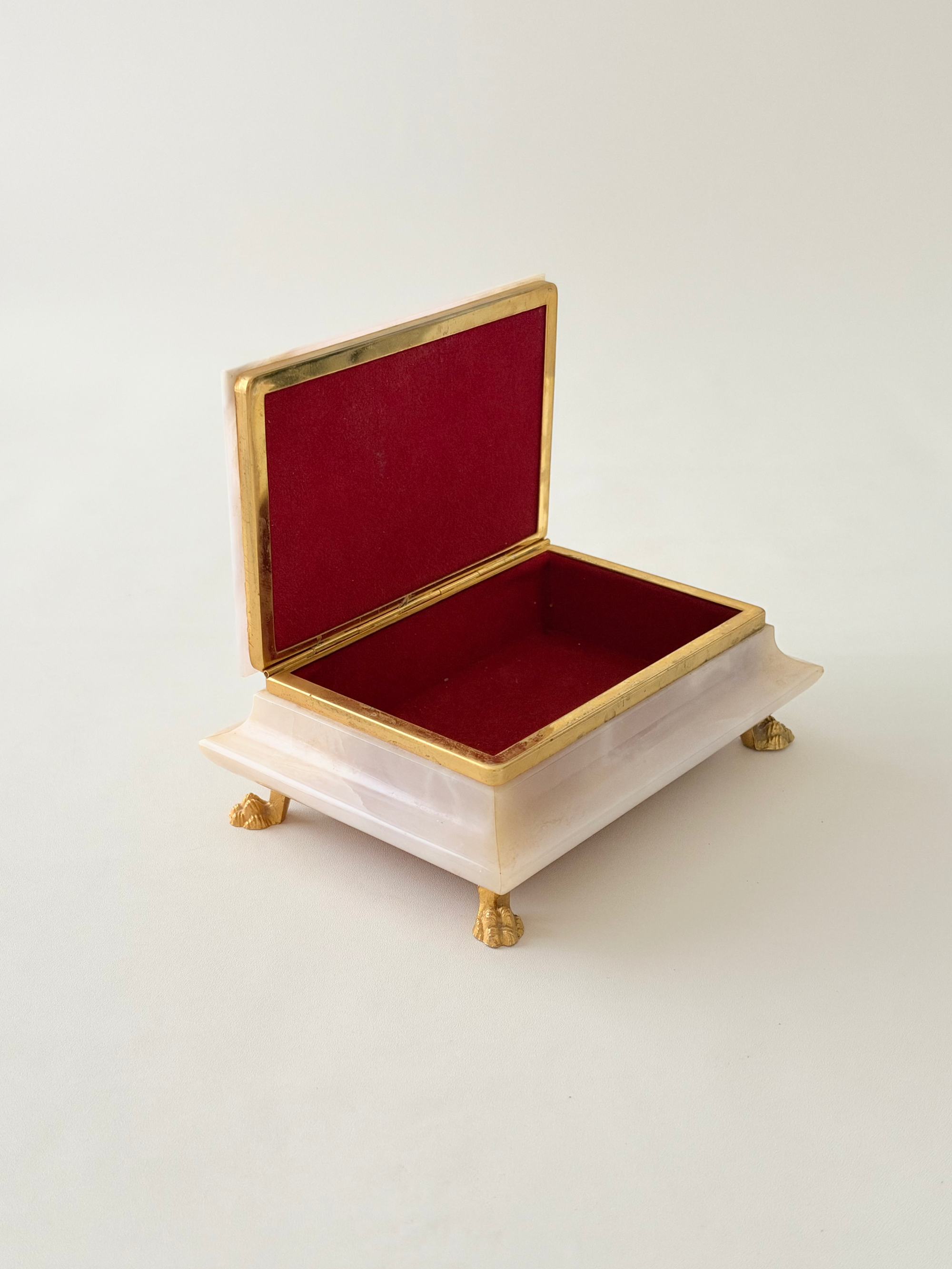 A dusty pink jewelry box with a  gold-tone metal trim, and a deep red felt interior, set against a plain off-white background.