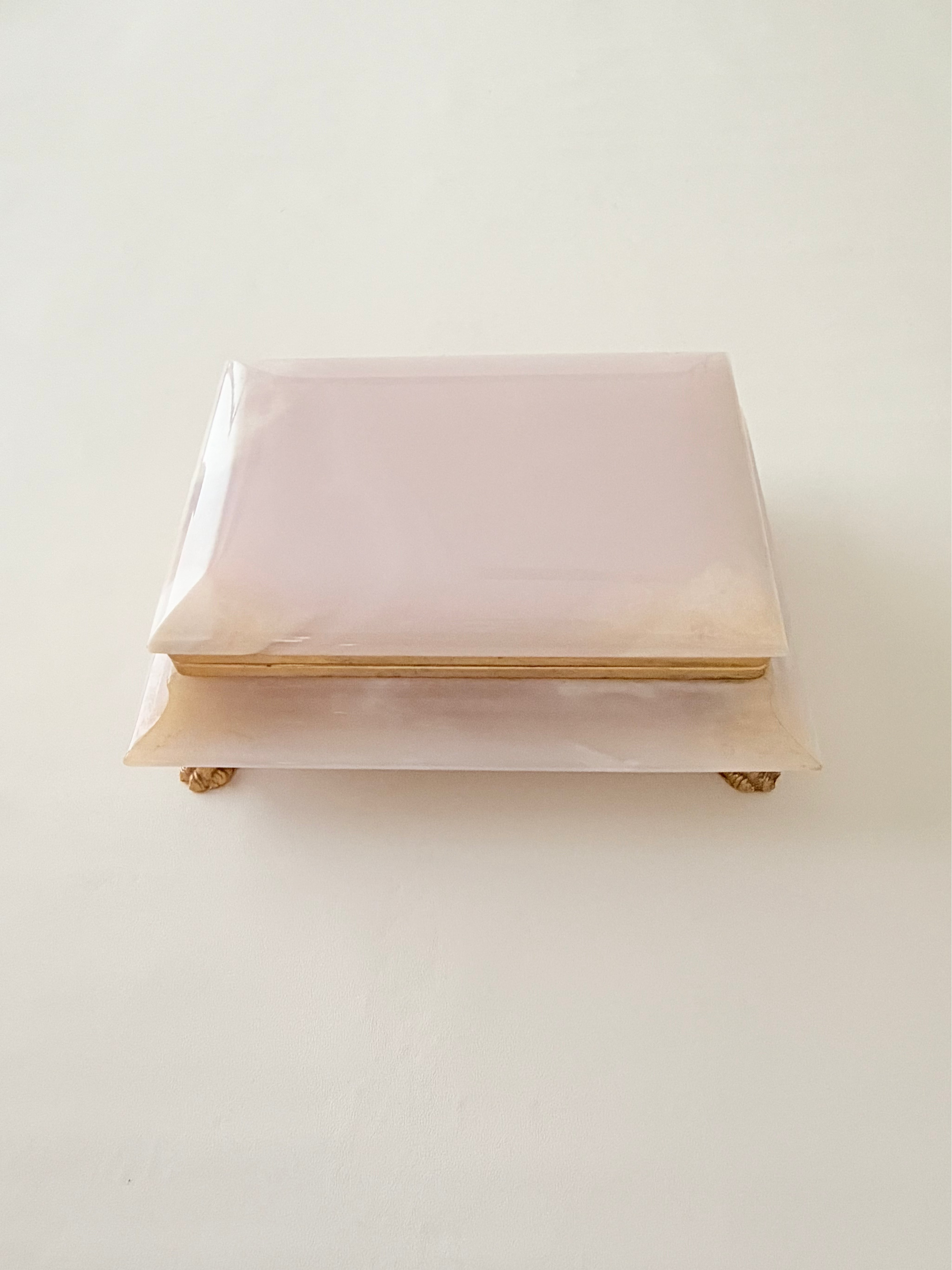 A rectangular dust pink  box with a hinged lid and gold-toned feet, photographed on a plain off-white background.