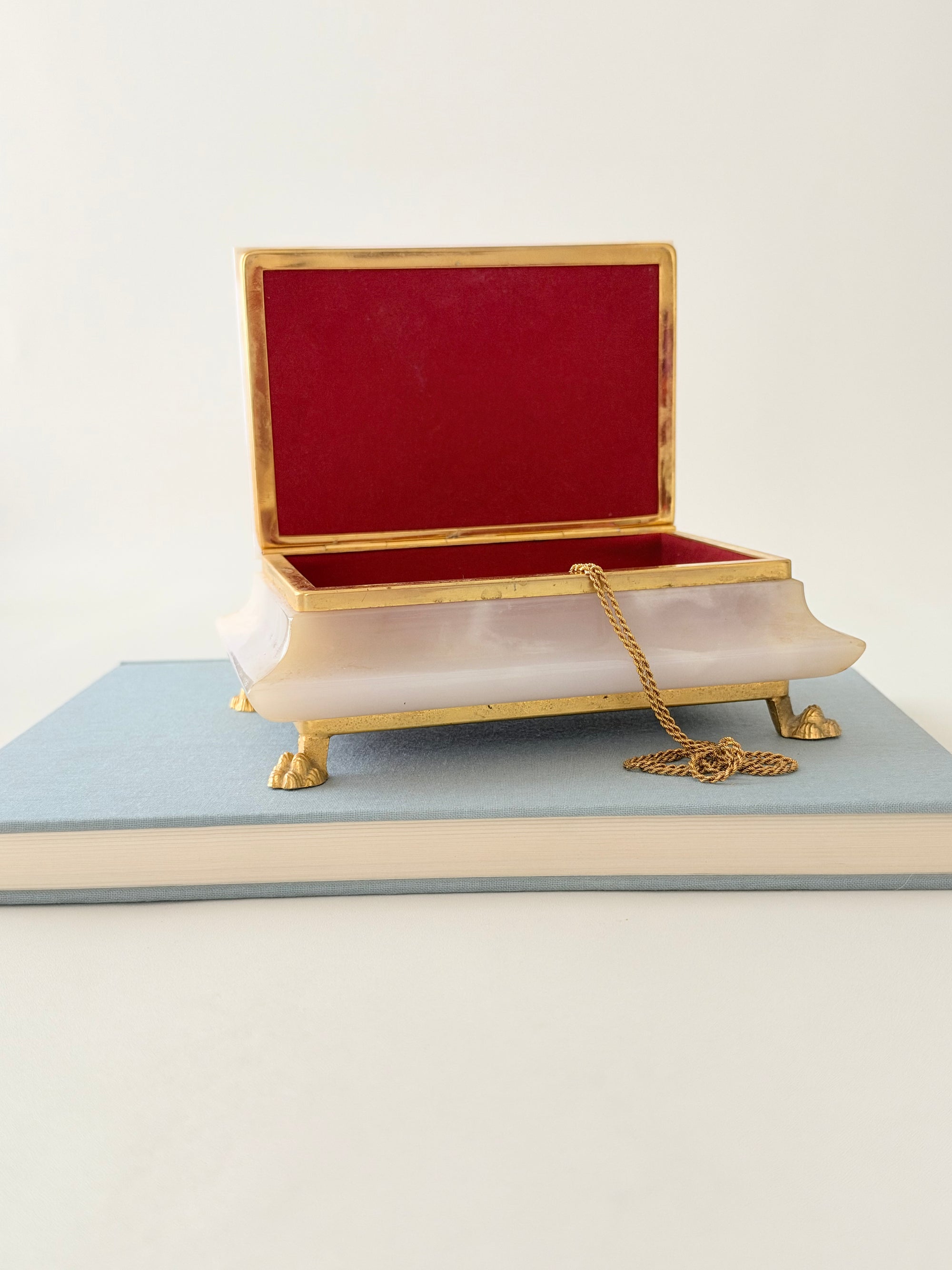 A dusty pink jewelry box gold-tone metal trim and feet, and a deep red felt-lined interior, shown open and resting on a light blue book against a white background.