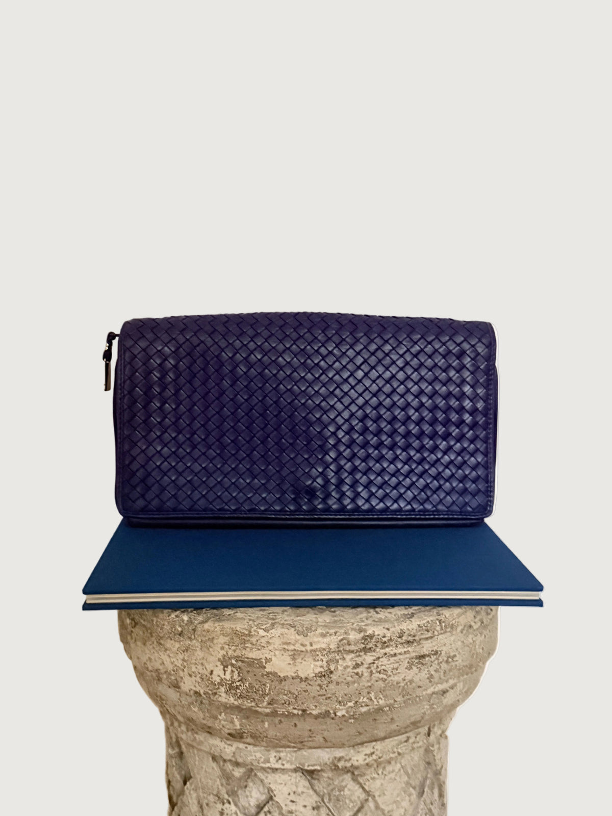 A purple woven leather clutch bag with a small black zipper pull, displayed on top of a blue book resting on a stone pedestal against a plain white wall.