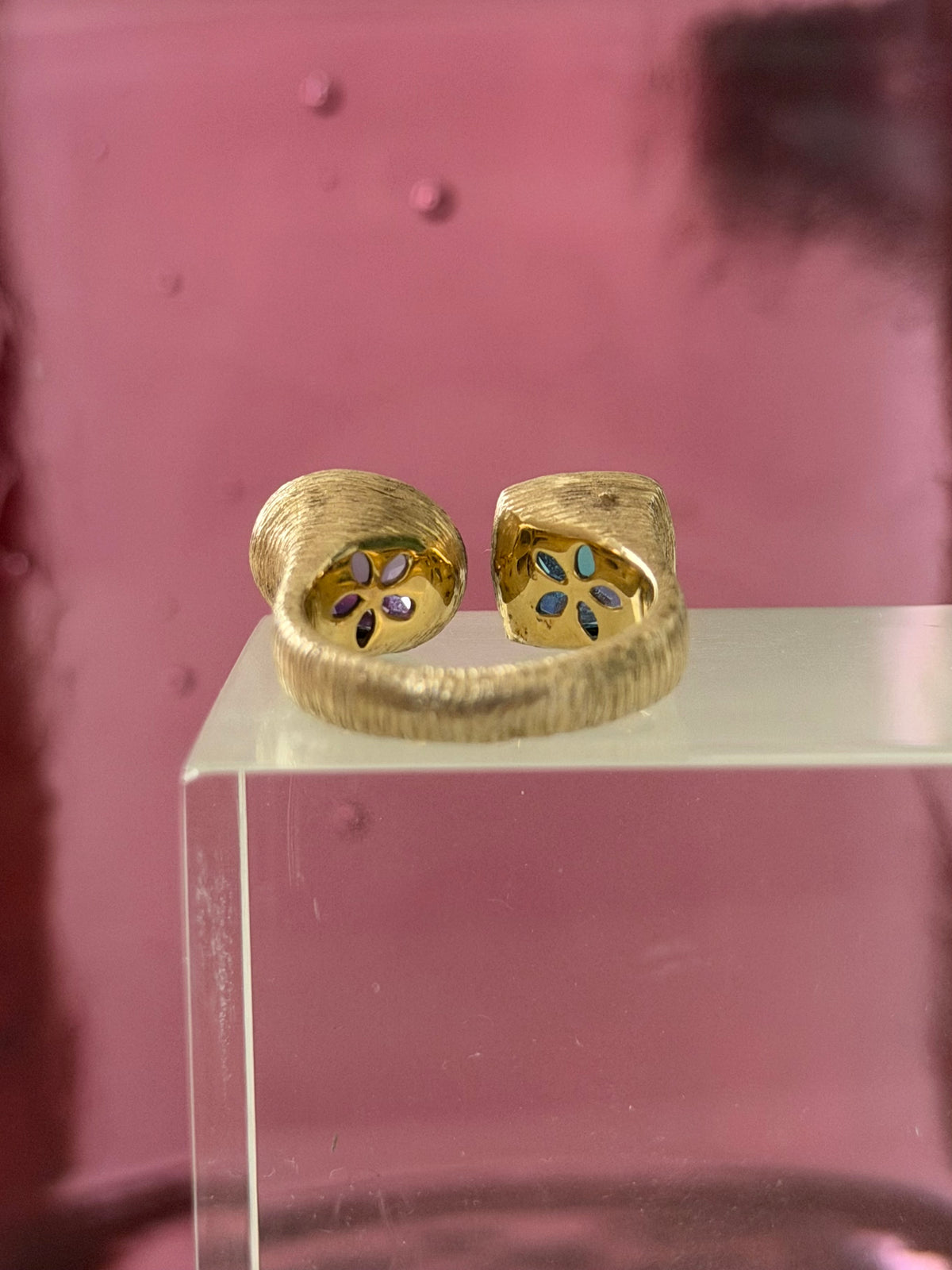 Inside view of a textured gold rings with blue and purple stones displayed on a clear acrylic block against a pink background.