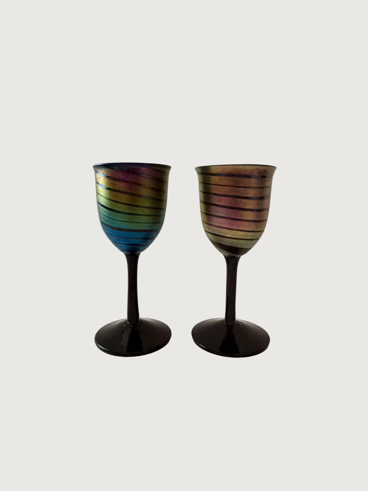 Two iridescent wine glasses with horizontal rainbow-colored stripes and dark stems, shown against a plain light gray background. Home Decor. Vintage. Collectible. 1970s.