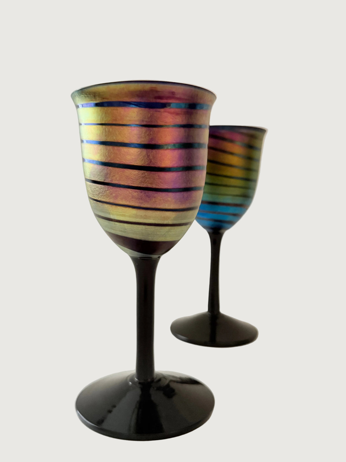 Two iridescent wine glasses with black stems and bases, featuring horizontal metallic stripes in gold, pink, blue, and purple, set against a light gray background. Home Decor. Vintage. Collectible. 1970s. 