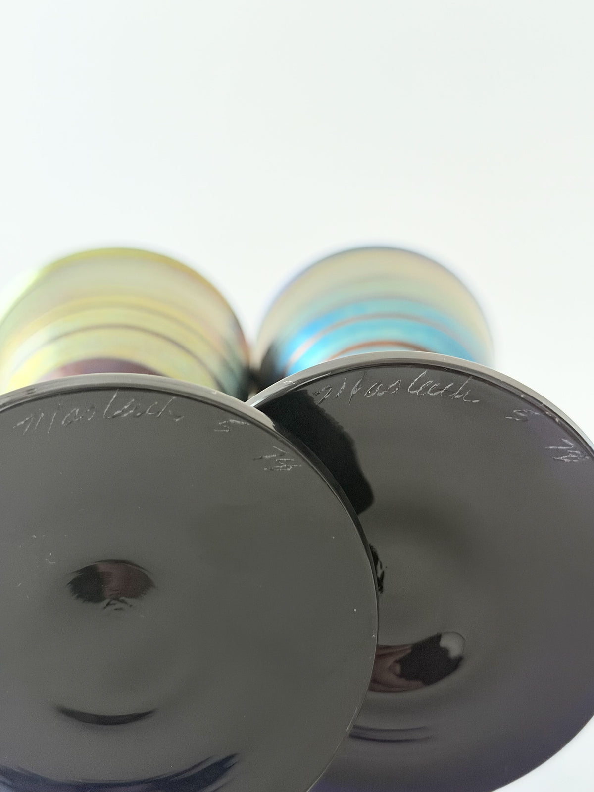 Signature Steven Maslach and date 1978 on two iridescent wine glasses with black stems and bases, featuring rainbow-colored horizontal stripes, displayed on top of a light blue book against a plain off-white background.  Home Decor. Vintage. Collectible. 1970s.