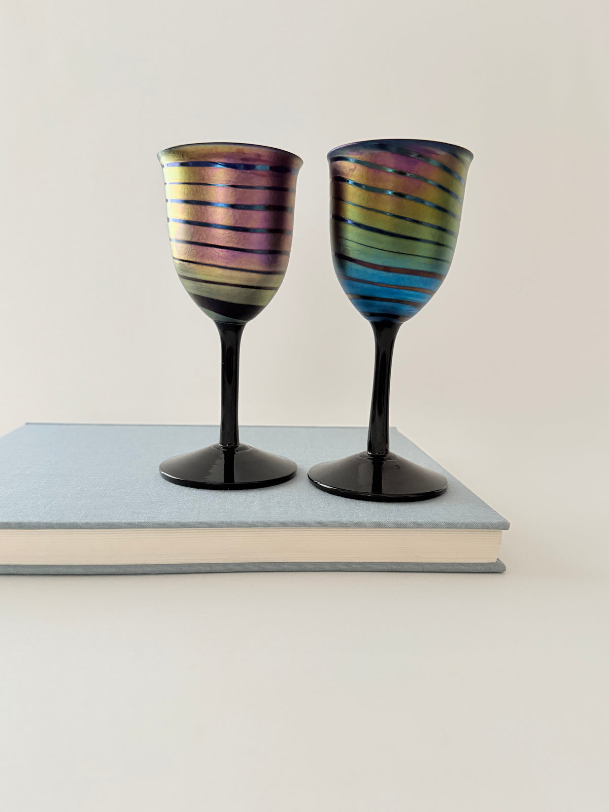 Two iridescent wine glasses with black stems and bases, featuring rainbow-colored horizontal stripes, displayed on top of a light blue book against a plain off-white background.  Home Decor. Vintage. Collectible. 1970s.