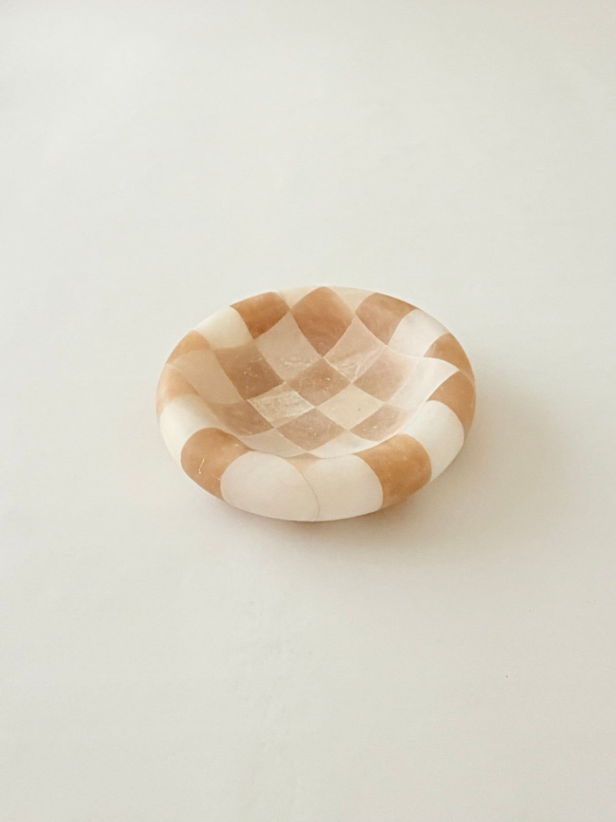 A small round decorative dish with a beige and off-white checkered pattern. Italian alabaster