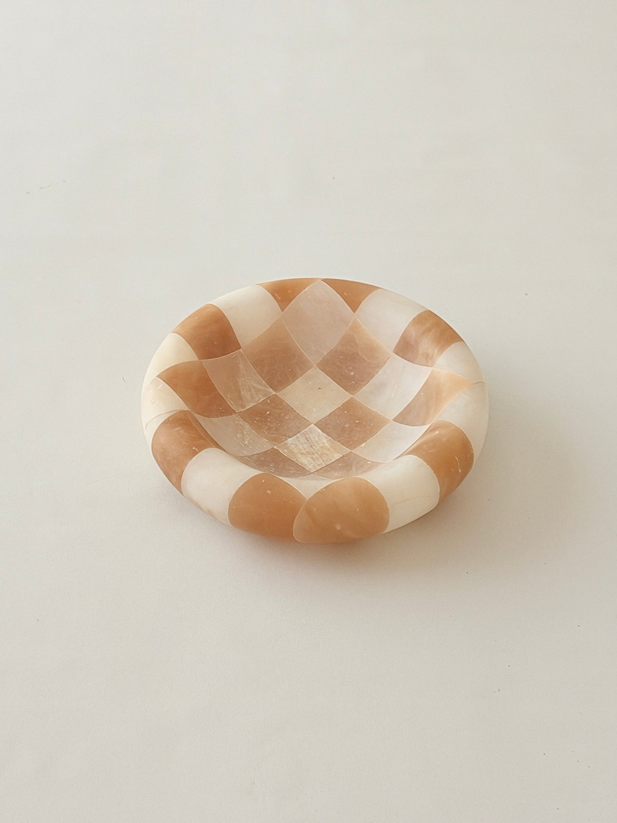 A small round dish with a beige and off-white checker patterned alabaster photographed on a plain light gray background.