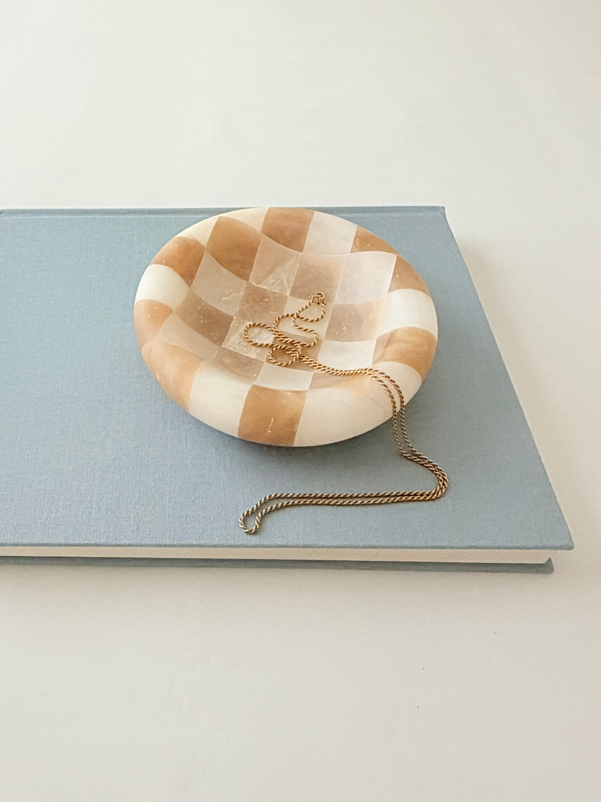 A round checkered alabaster jewelry alabaster tray styled on top of a light blue hardcover book with a gold chain resting inside the tray.