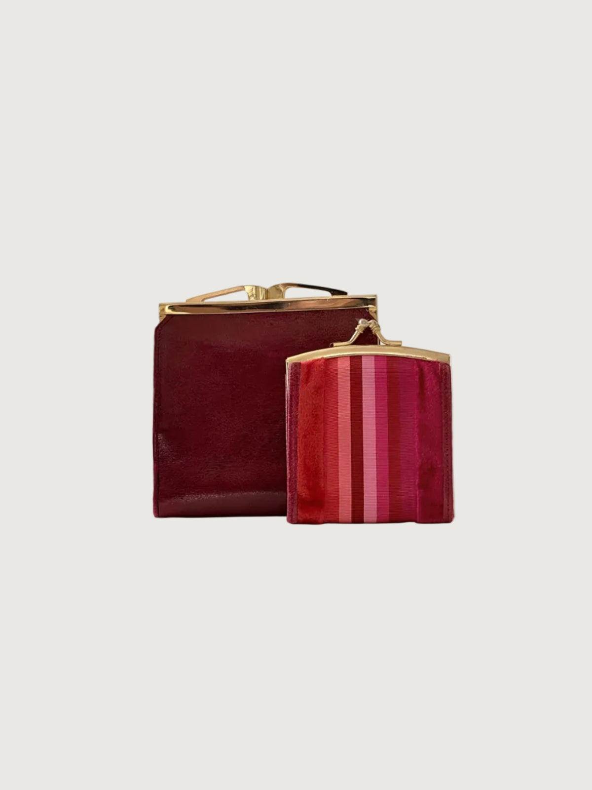 Two small wallets with gold-tone metal frames on a light gray studio background. The larger clutch behind is deep burgundy, and the smaller front clutch has vertical pink and red stripes.