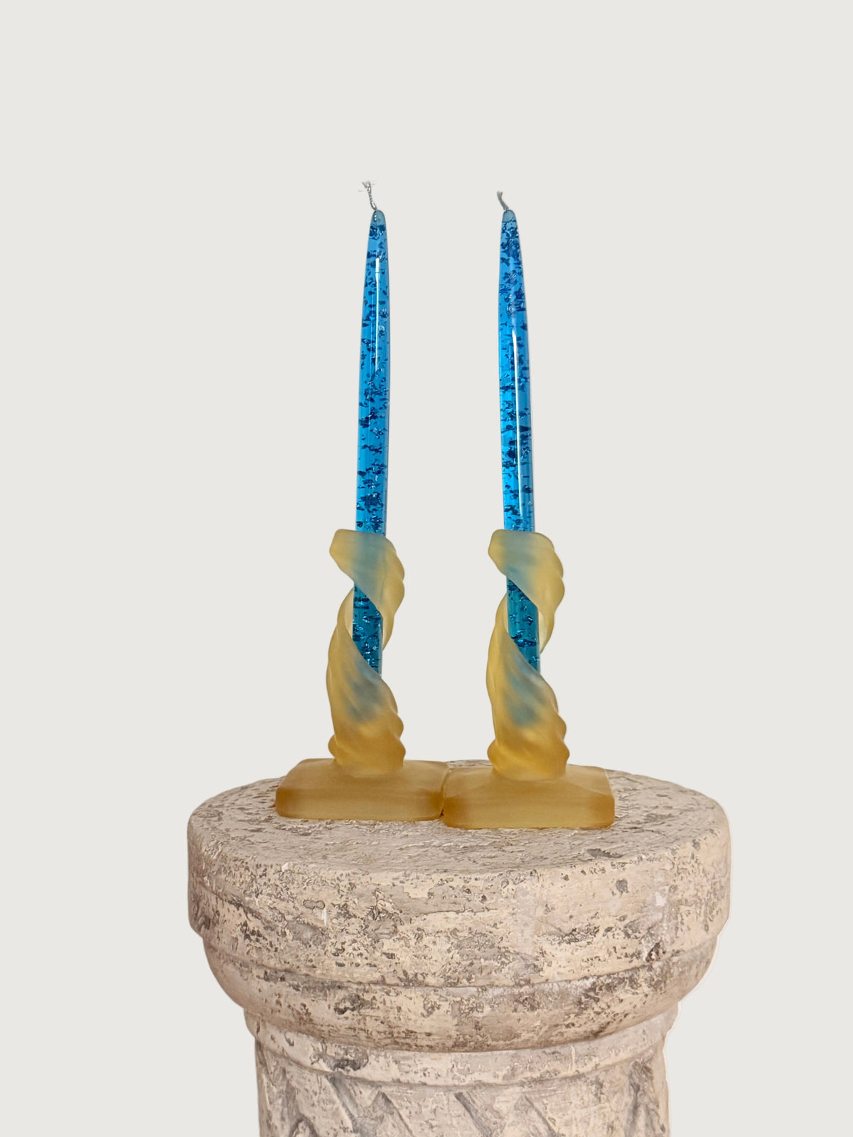 A pair of tall blue lucite candles in a  pair of satin pressed glass yellow twisted glass candleholders, displayed on a stone pedestal against a plain off-white wall.